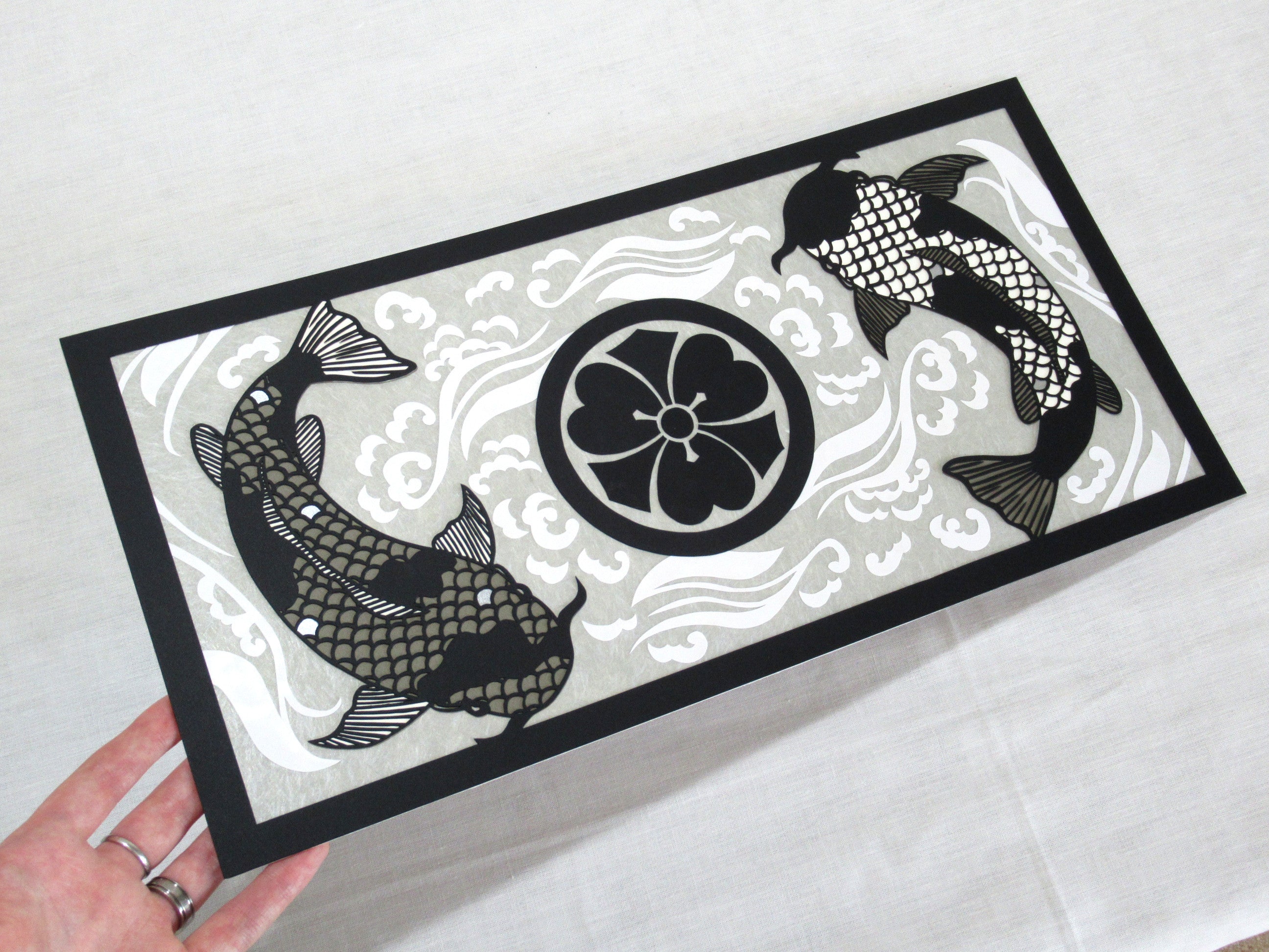 Image of an intricate cut paper piece, with two black and grey koi circling a black Japanese family crest all on a grey background with white swirling waves.