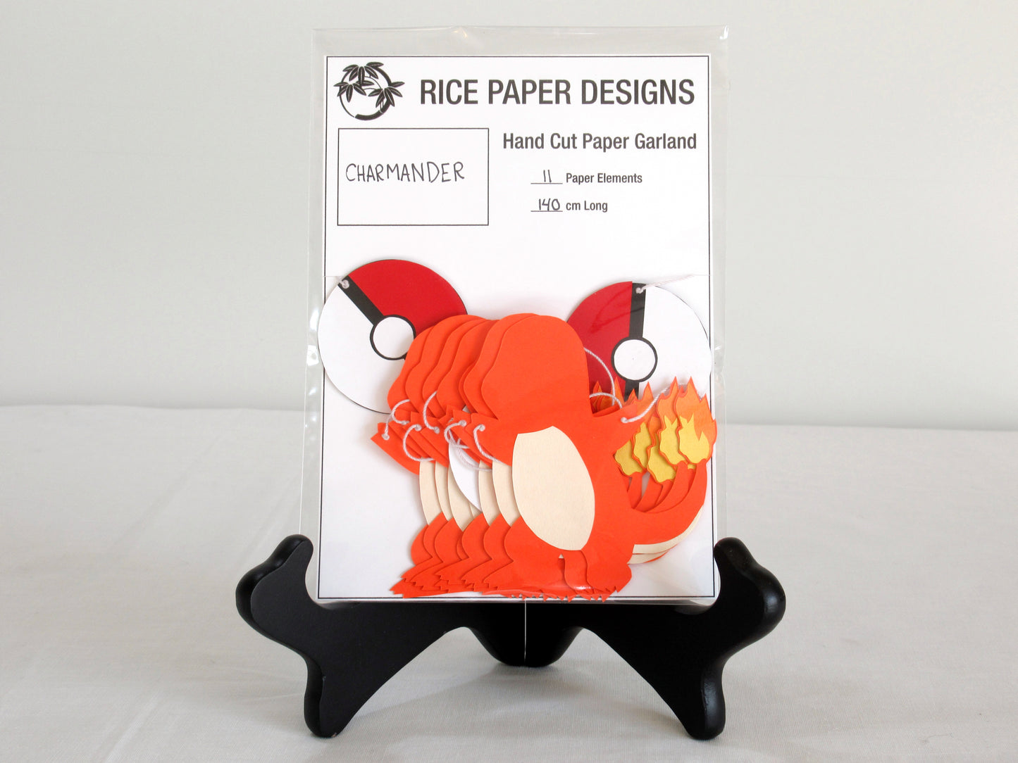 A garland with a series of paper charmanders and pokeballs arranged in neat bundle inside a clear bag. There is a paper behind them showing the Rice Paper Designs logo, and information about the garland.