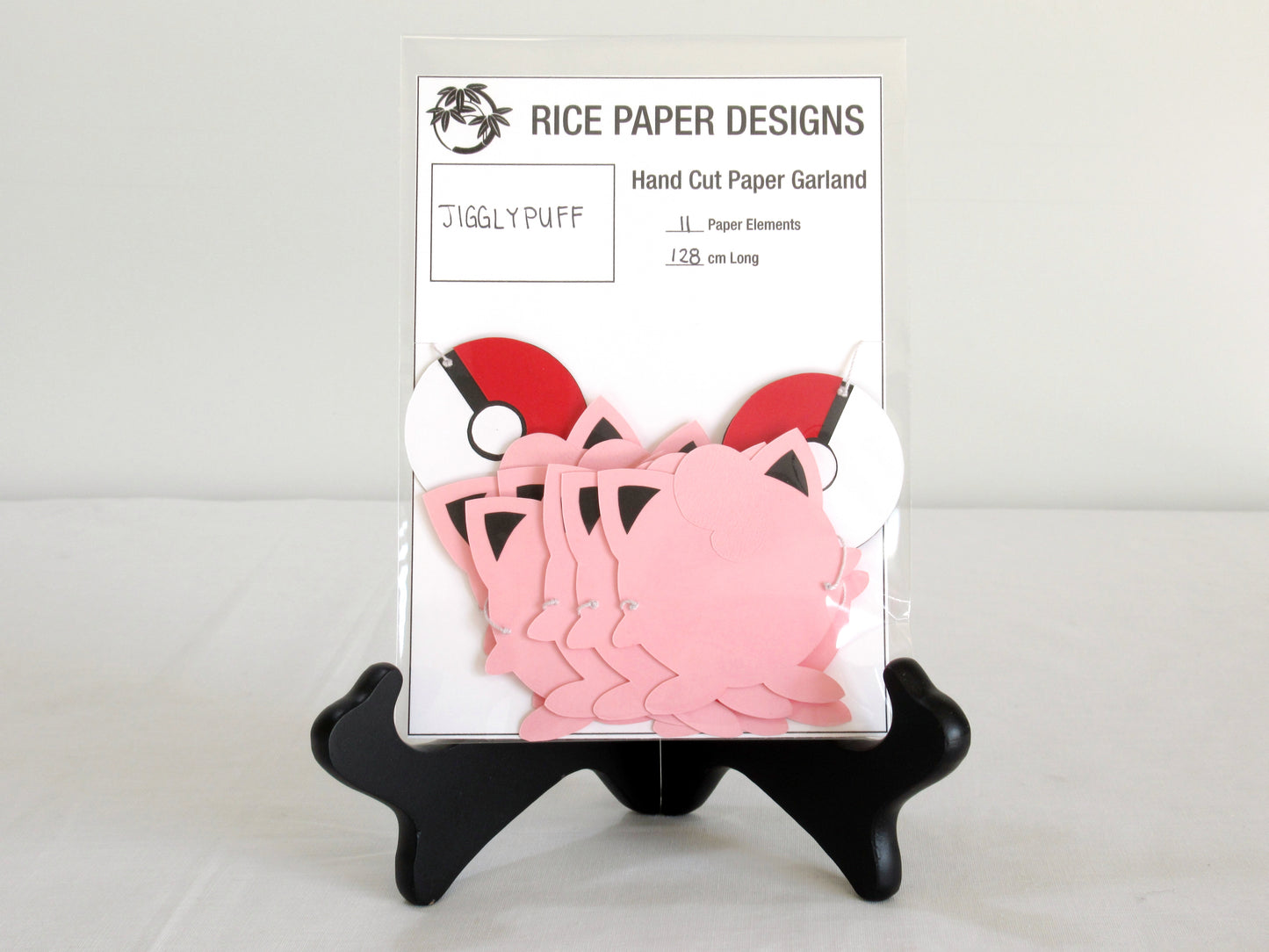 A garland with a series of paper jigglypuffs and pokeballs arranged in neat bundle inside a clear bag. There is a paper behind them showing the Rice Paper Designs logo, and information about the garland.