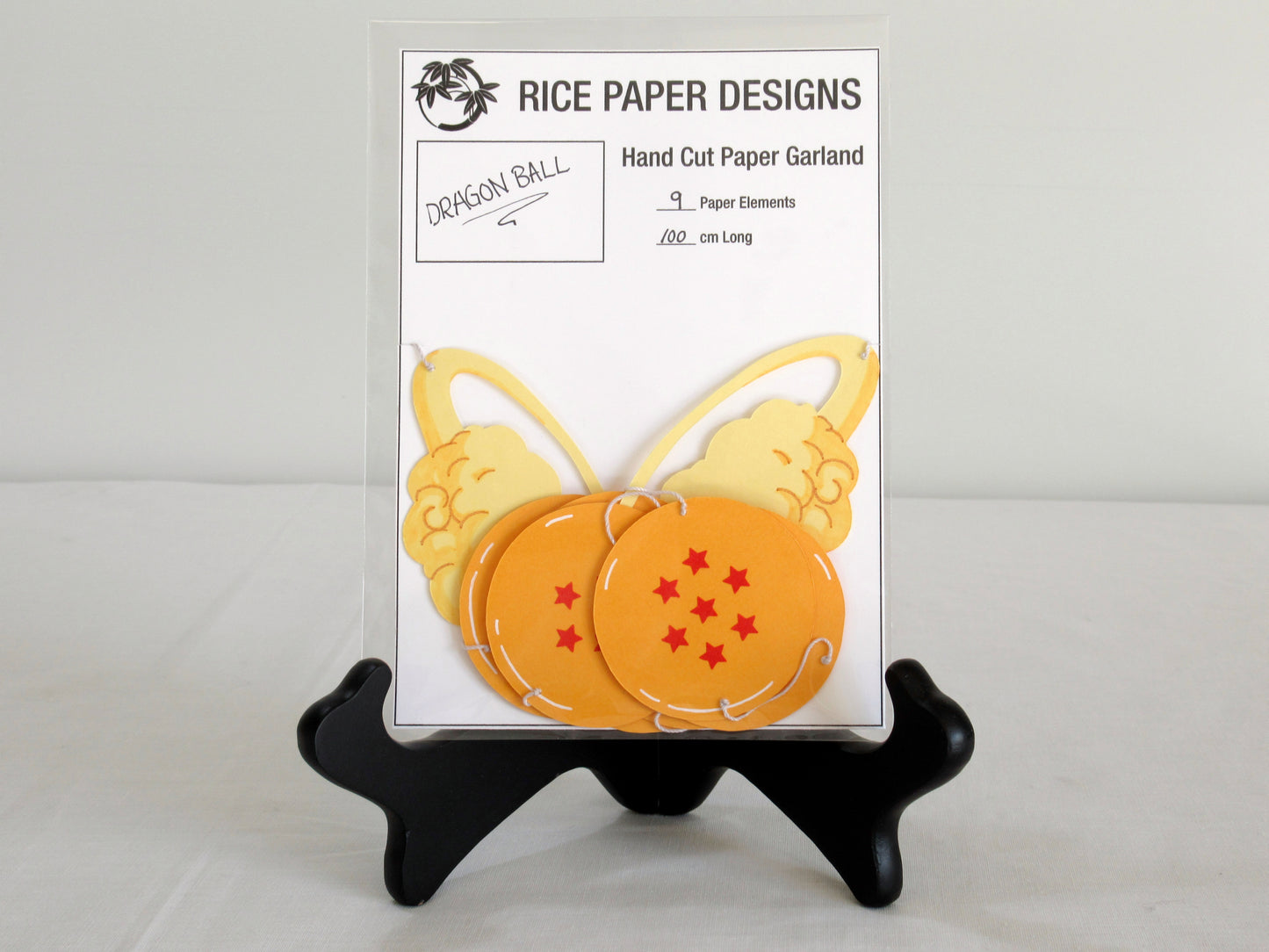 A garland with a series of paper dragon balls and nimbus arranged in neat bundle inside a clear bag. There is a paper behind them showing the Rice Paper Designs logo, and information about the garland.