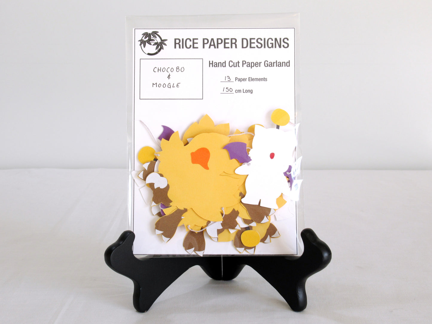 A garland with a series of paper chocobo and moogles arranged in neat bundle inside a clear bag. There is a paper behind them showing the Rice Paper Designs logo, and information about the garland.