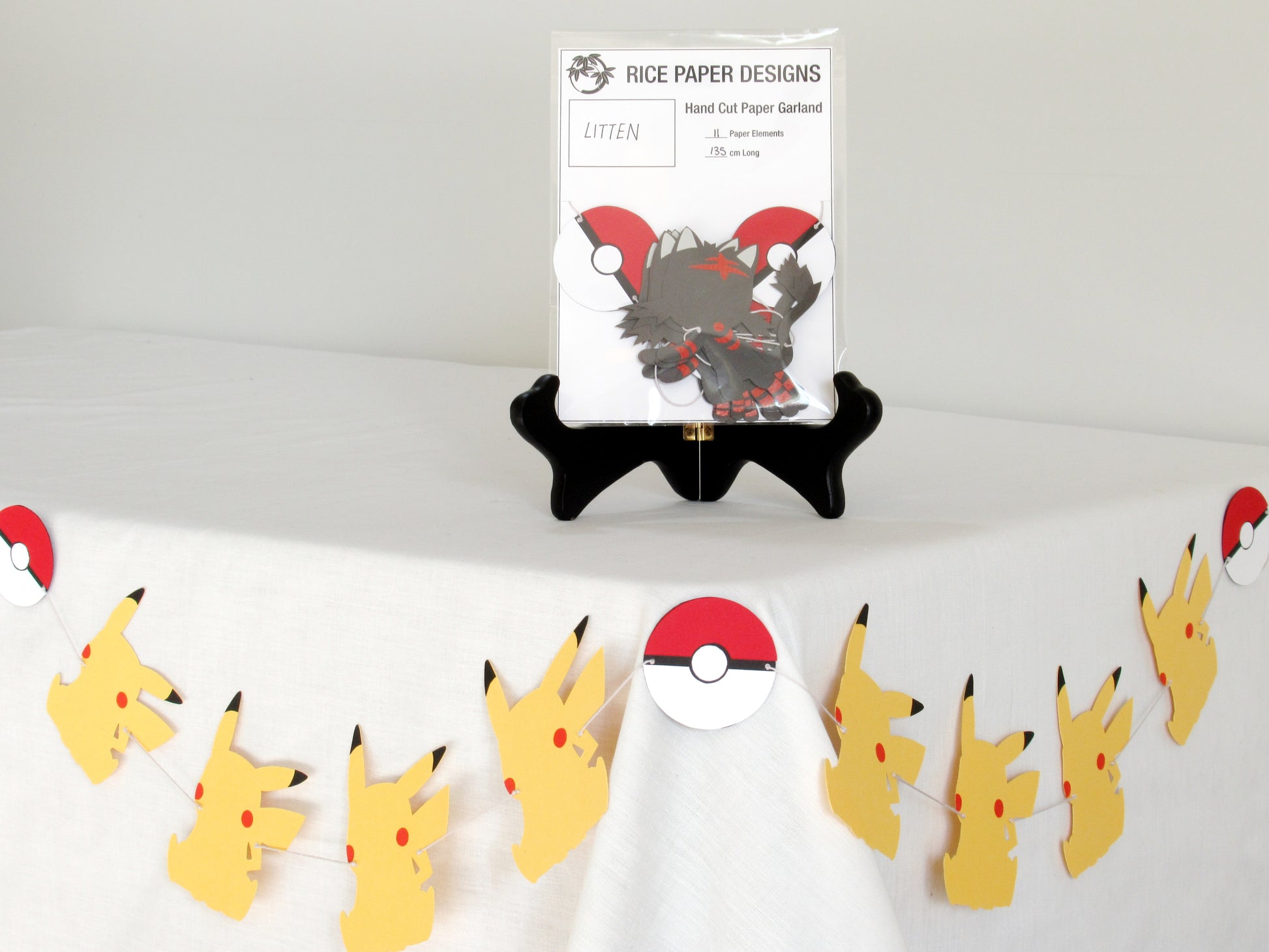 A garland with a series of paper littens and pokeballs arranged in neat bundle inside a clear bag. There is a paper behind them showing the Rice Paper Designs logo, and information about the garland. The bag is on a table, and below it is a sample garland with pikachu hung up.
