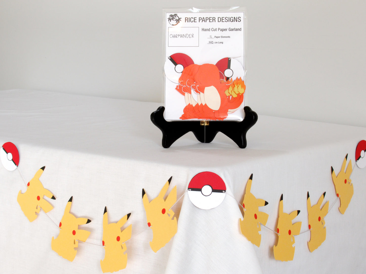 A garland with a series of paper charmanders and pokeballs arranged in neat bundle inside a clear bag. There is a paper behind them showing the Rice Paper Designs logo, and information about the garland. The bag is on a table, and below it is a sample garland with pikachu hung up.