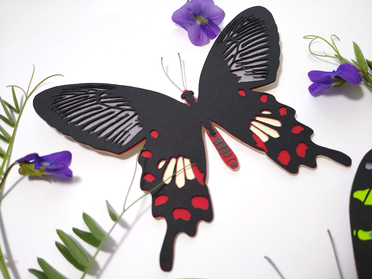 Multi-layered paper butterfly, in the design of a Common Rose butterfly. It rests next to several sprigs of leaves and small purple flowers.