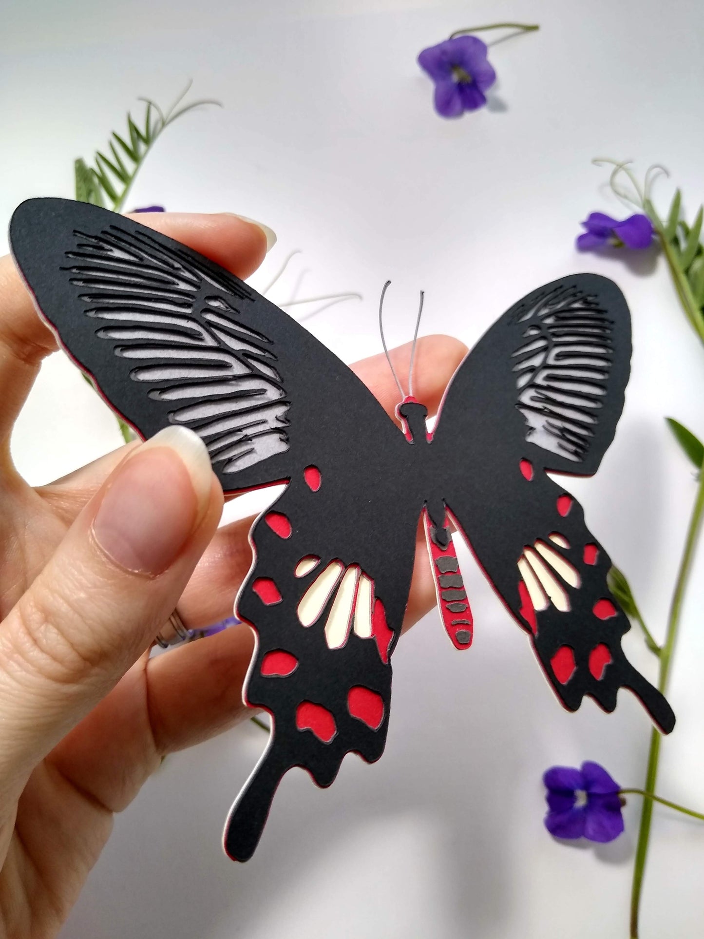 A hand holds a multi-layered paper butterfly to the camera, in the design of a Common Rose butterfly. In the background are several sprigs of leaves and small purple flowers.