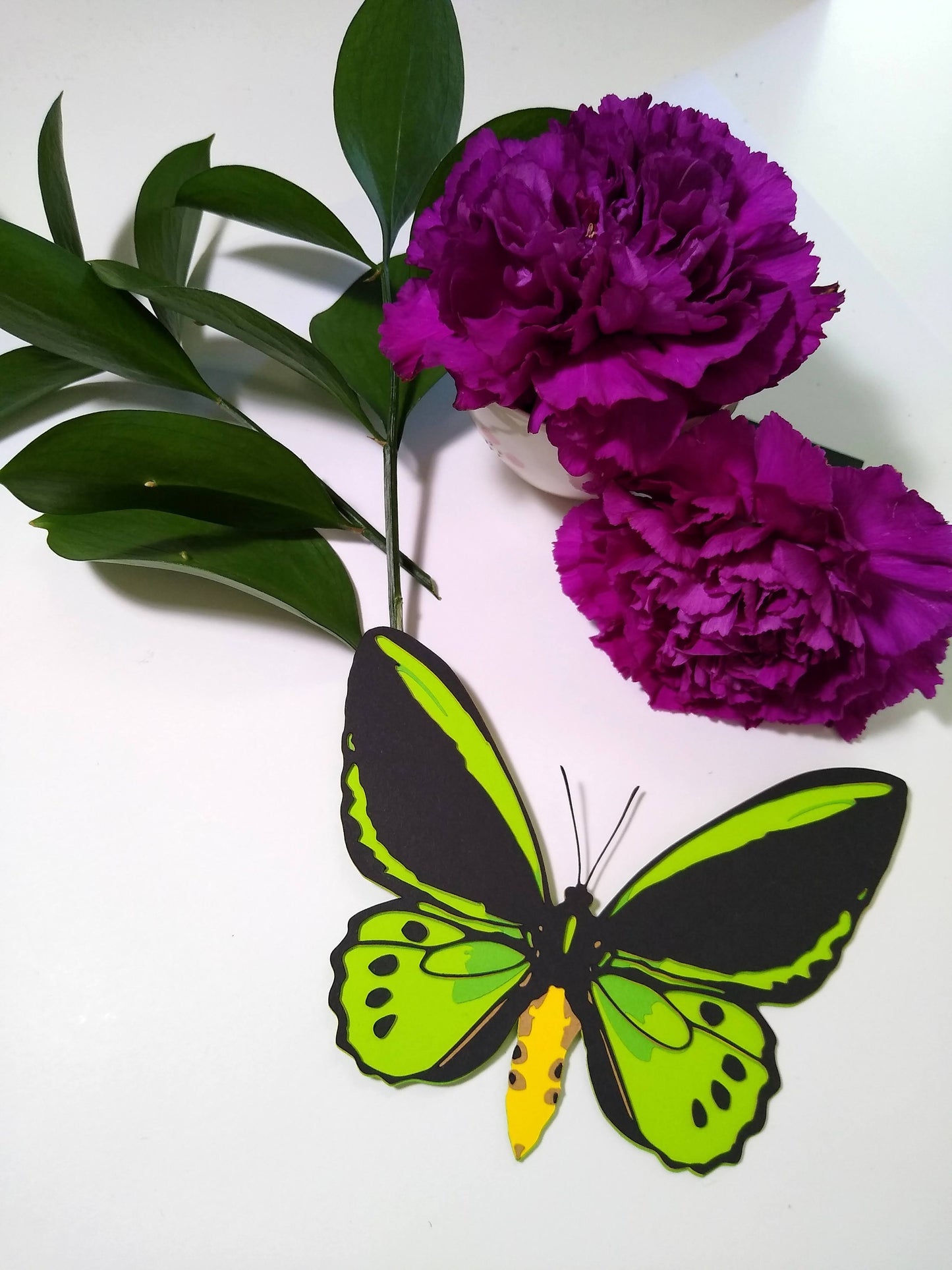 A multi-layered paper butterfly rests on a table. It is in the design of the front of a Cairns Birdwing butterfly. It rests below sprigs of leaves and two purple flowers.