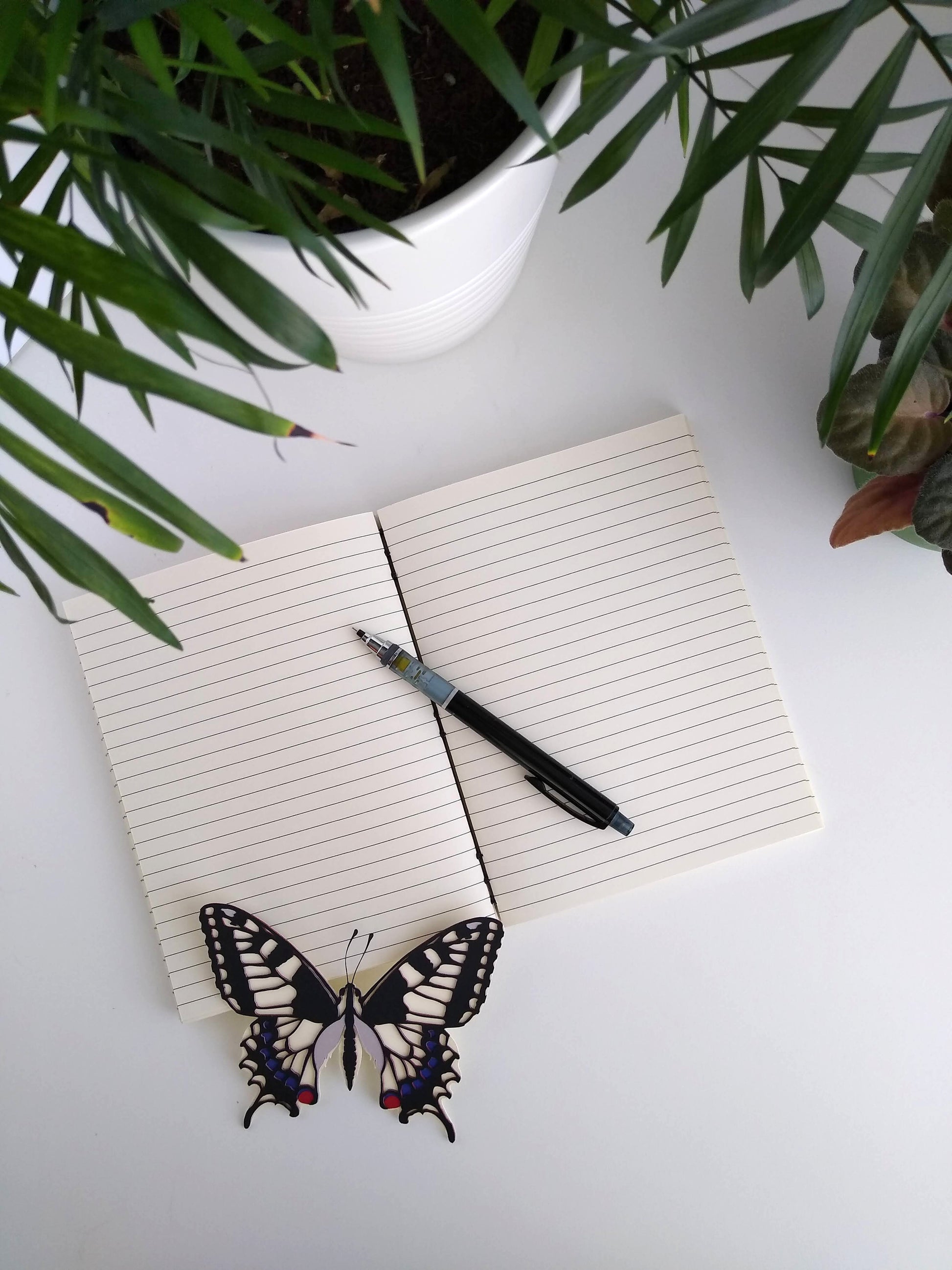 An open journal lays on a white desk next to two potted plants and a paper butterfly. The journal has a grey thread connecting the pages at the spine and a black mechanical pencil rests across the lined, cream pages.