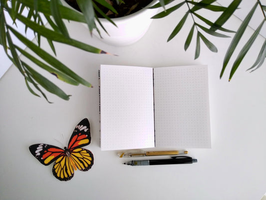 Handmade journal, opened up flat showing pages with a dotted grid on it. It's laying on a desk next to a potted plant, a gold pen and a black mechanical pencil, and paper butterfly.