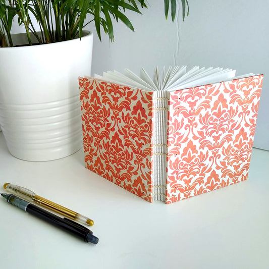 A handmade journal sits upright beside a potted plant, a gold pen, and a black mechanical pencil. The cover of the journal is cream with an orange damask pattern printed across it. It's angled to show the open spine and the cream stitching holding it together.