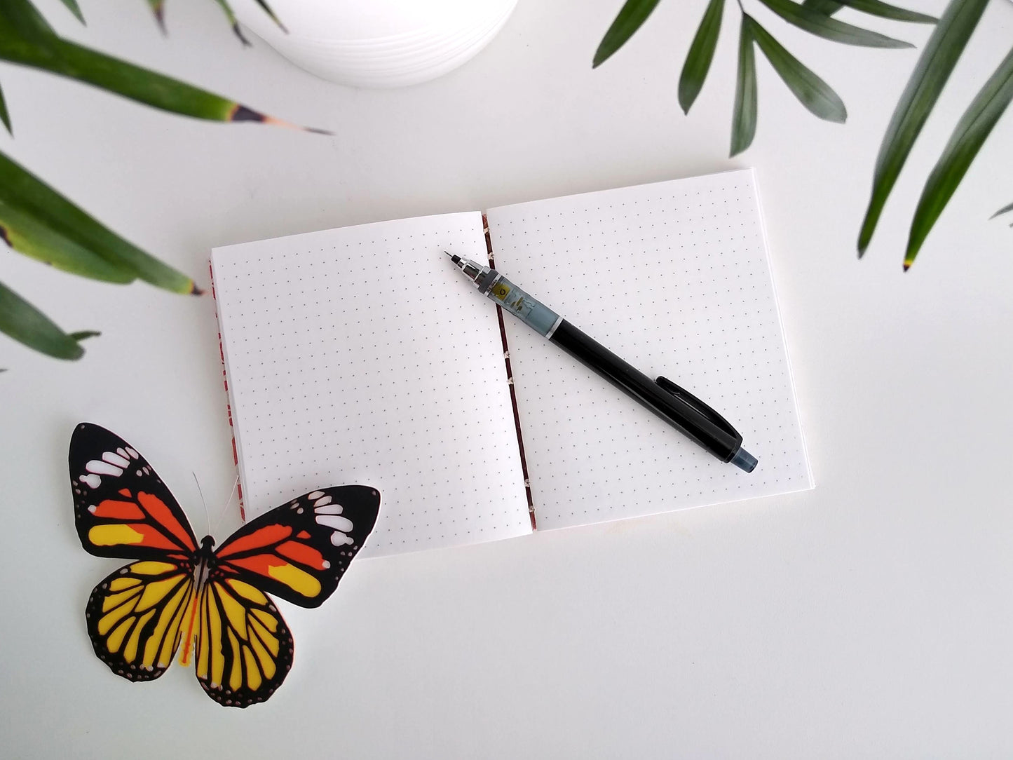 An open journal lays on a white desk next to a potted plant and a paper butterfly. The journal has a cream thread connecting the pages at the spine and a black mechanical pencil rests across the white pages printed with a dot grid.