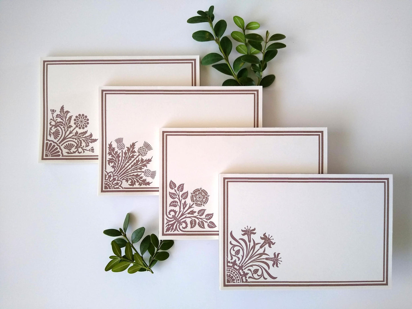 Four cream colored notepads on a white background. Sprigs of leaves rest around them. Each notepad has a stylized floral design in tan in the lower left corner with a double line border around the rest of the notepad.