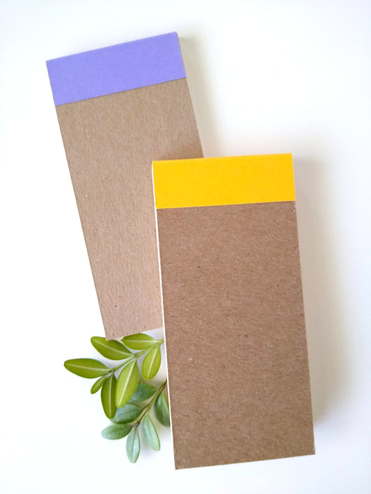 The back of the notepads shows how the colored covers are glued to the back cardboard. They rest on a white background with two sprigs of green leaves around them.