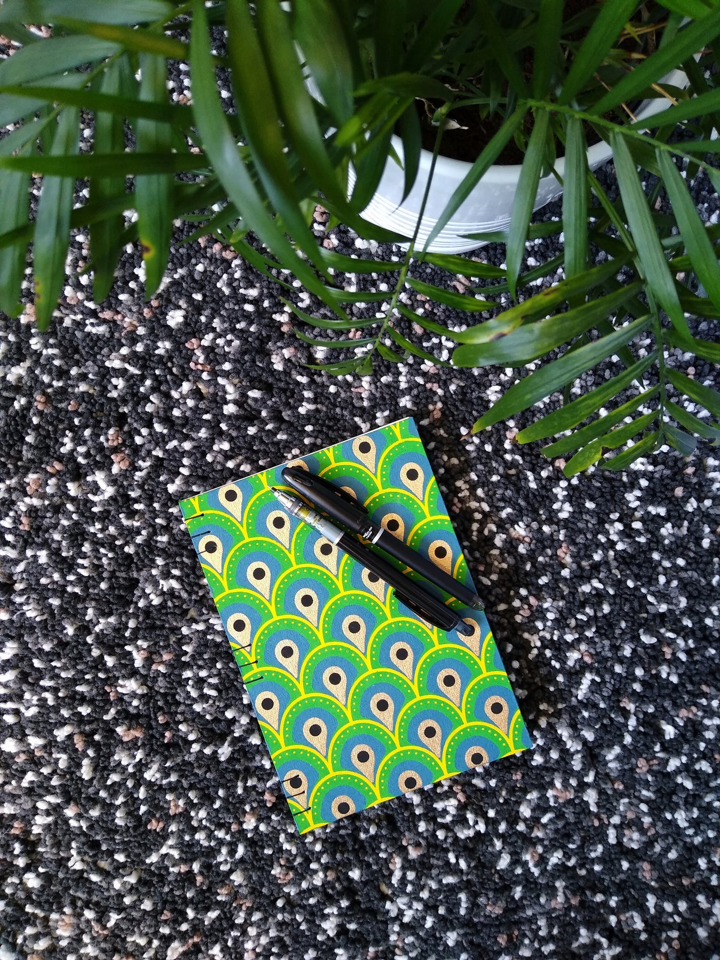 A handmade journal laying on a black shag carpet beside a potted plant, with a black pen and mechanical pencil on top. The cover of the journal is an abstract peacock feather design.