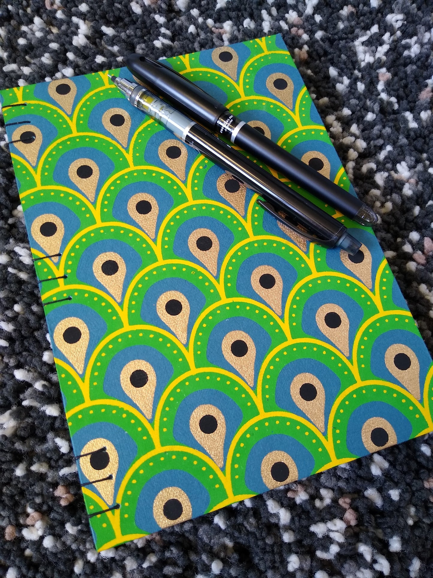 A close up of handmade journal laying on a black shag carpet with a black pen and mechanical pencil on top. The cover of the journal is an abstract peacock feather design.
