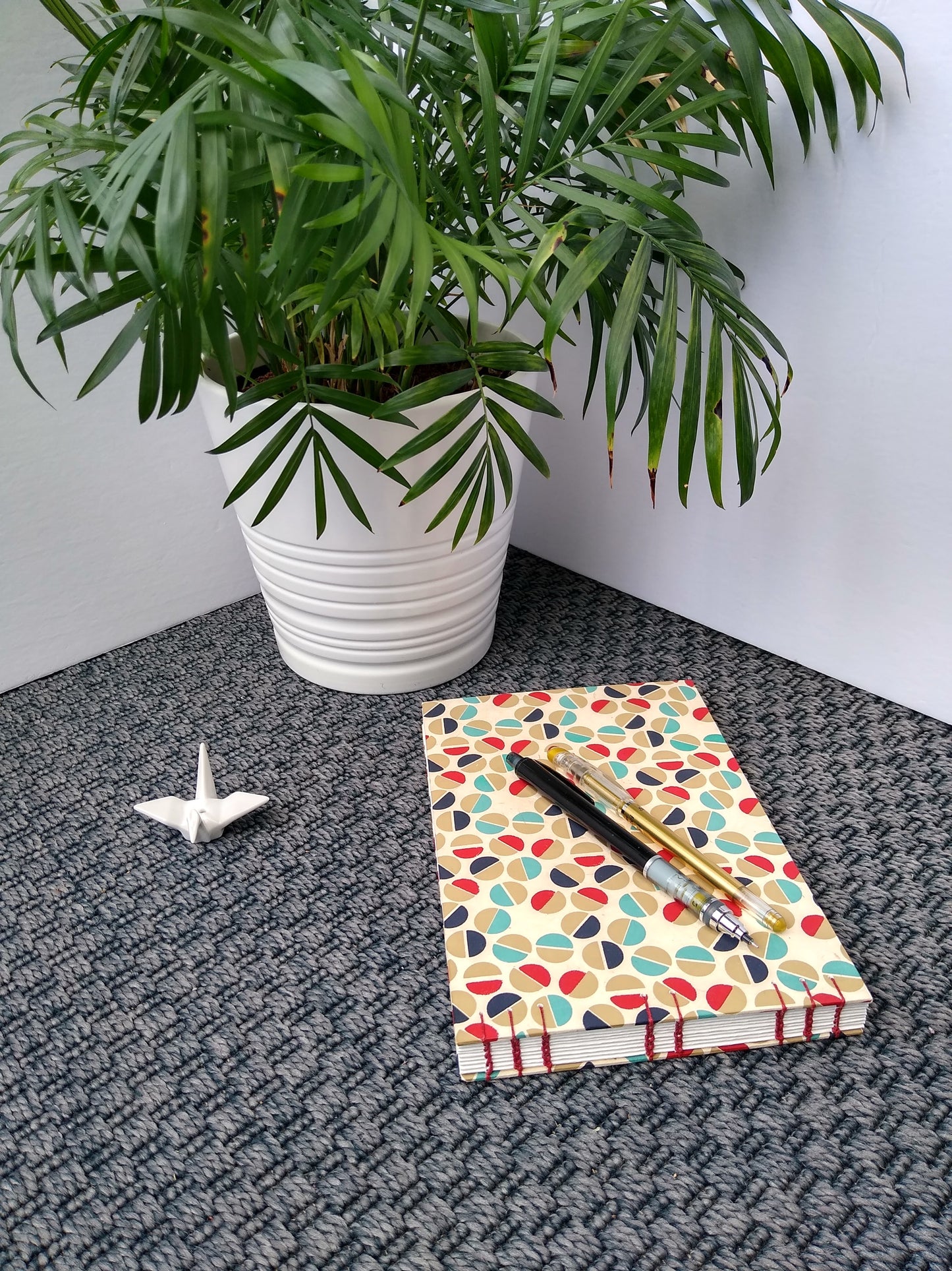 A handmade journal laying on a grey carpet beside a potted plant, a ceramic crane, a gold pen, and a black mechanical pencil. The cover of the journal is cream with half circles in gold, teal, dark blue, and red all over it. The half circles are placed next to each other to look like the head of a flat-head screw. The spine is facing the camera so the view can see the red thread stitching holding the journal together.
