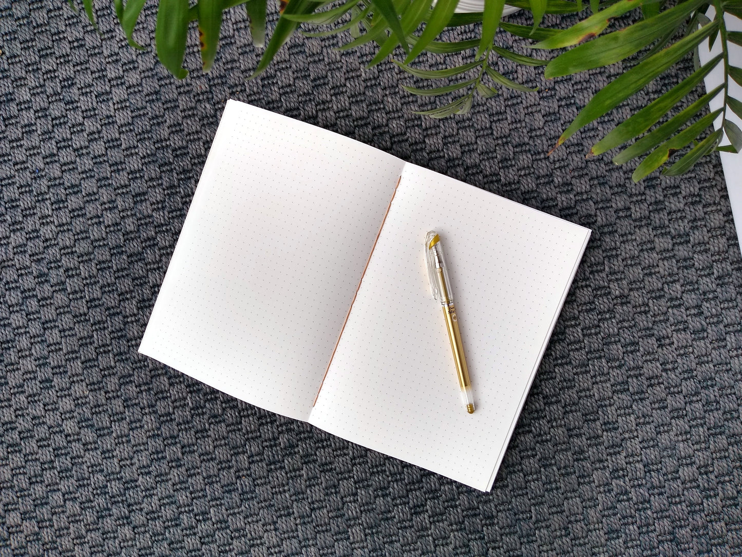 An open journal lays on a grey carpet next to a potted plant. The journal has a brown thread connecting the pages at the spine and a gold pen rests across the pages that have a dotted grid printed on them.