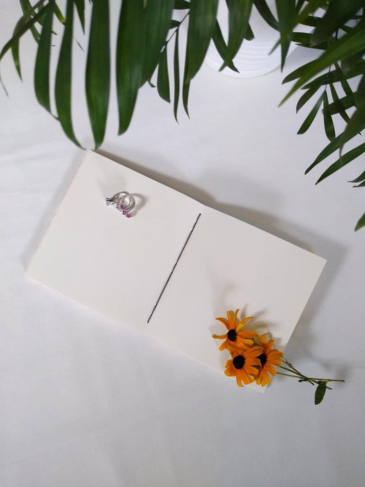 An open journal lays on a white desk next to a potted plant. The journal has a black thread connecting the pages at the spine and two rings and three yellow flowers resting across the blank white pages.