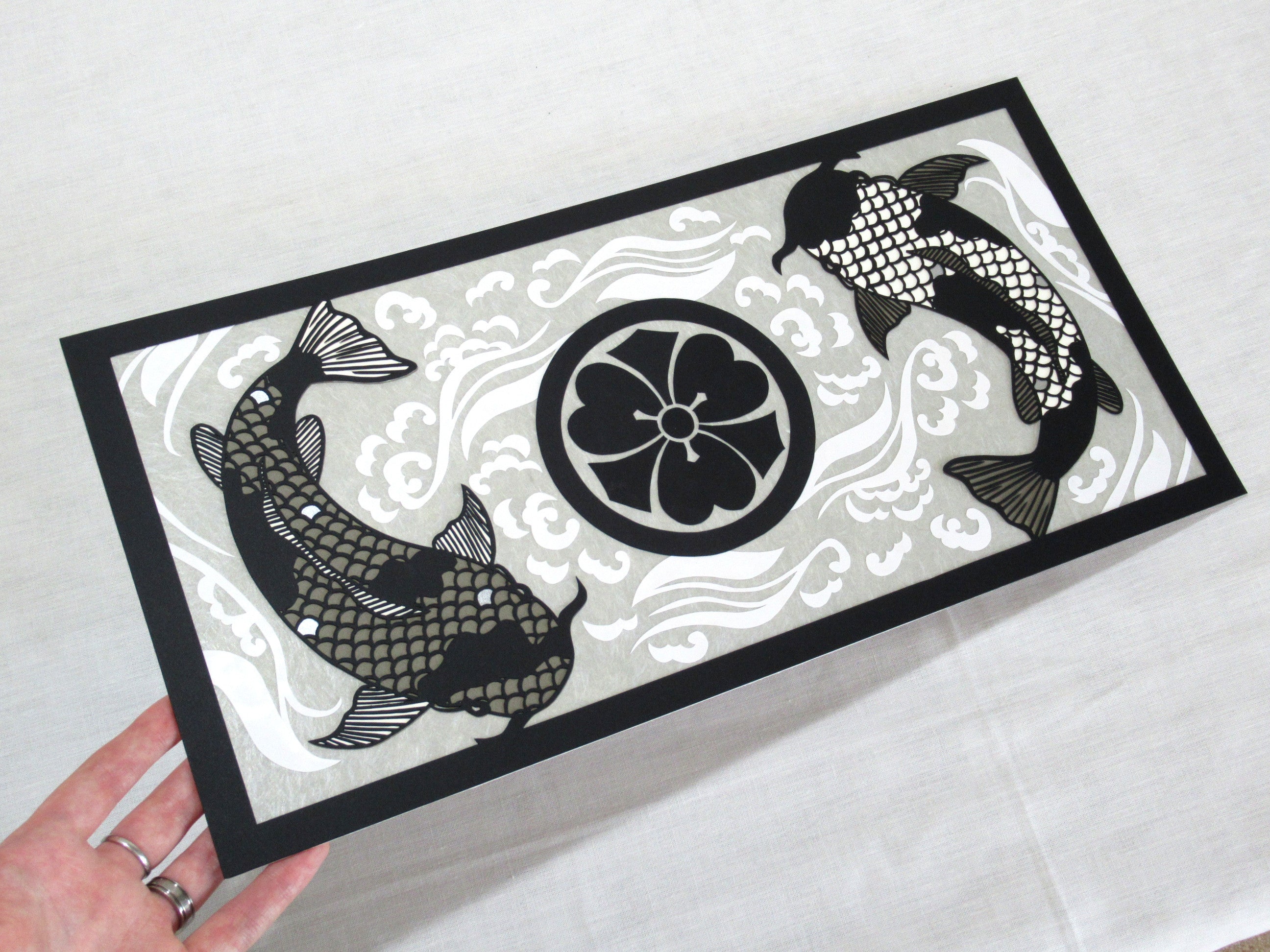 Image of an intricate cut paper piece, with two black and grey koi circling a black Japanese family crest all on a grey background with white swirling waves. 