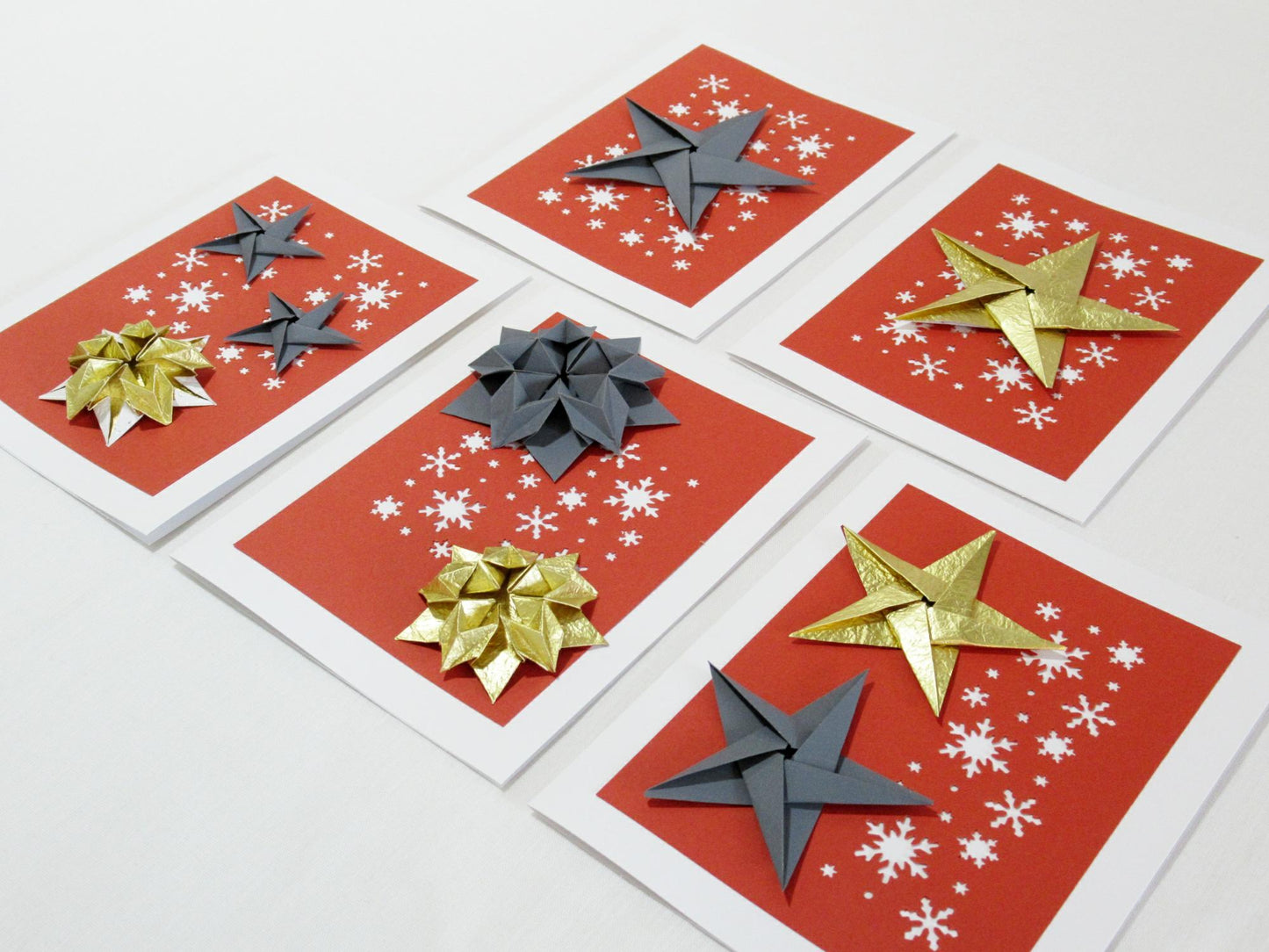 Set of five white cards. Each card has a red background with white snowflakes. On top are a variety of origami stars in both gold and grey.