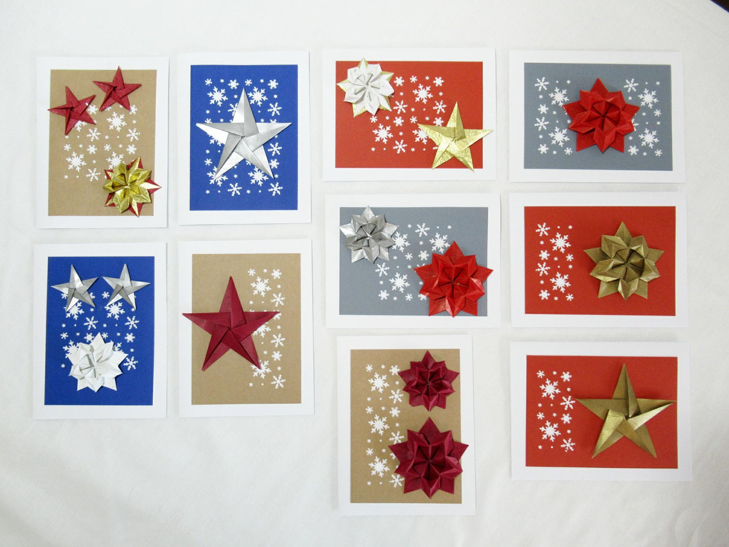 Set of ten white cards. Each card has either a red, blue, grey, or kraft background with white snowflakes. On top are a variety of origami stars in red, silver, white, and gold.