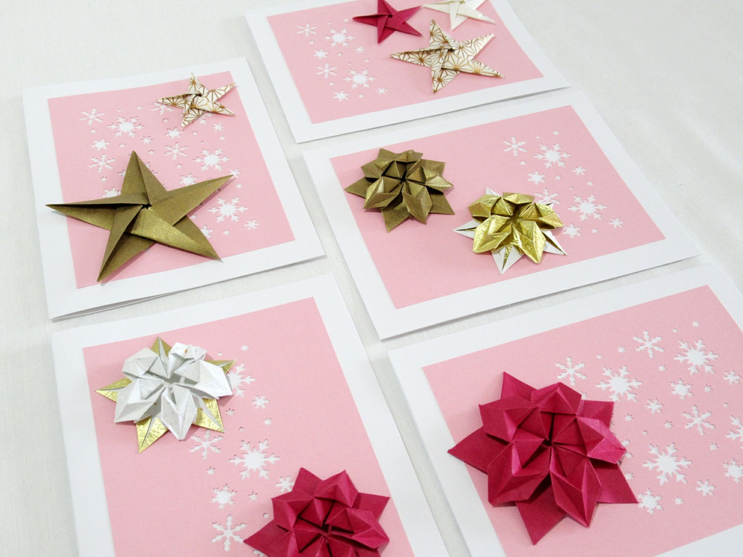 Set of five white cards. Each card has a pink background with white snowflakes. On top are a variety of origami stars in gold, white, and pink.