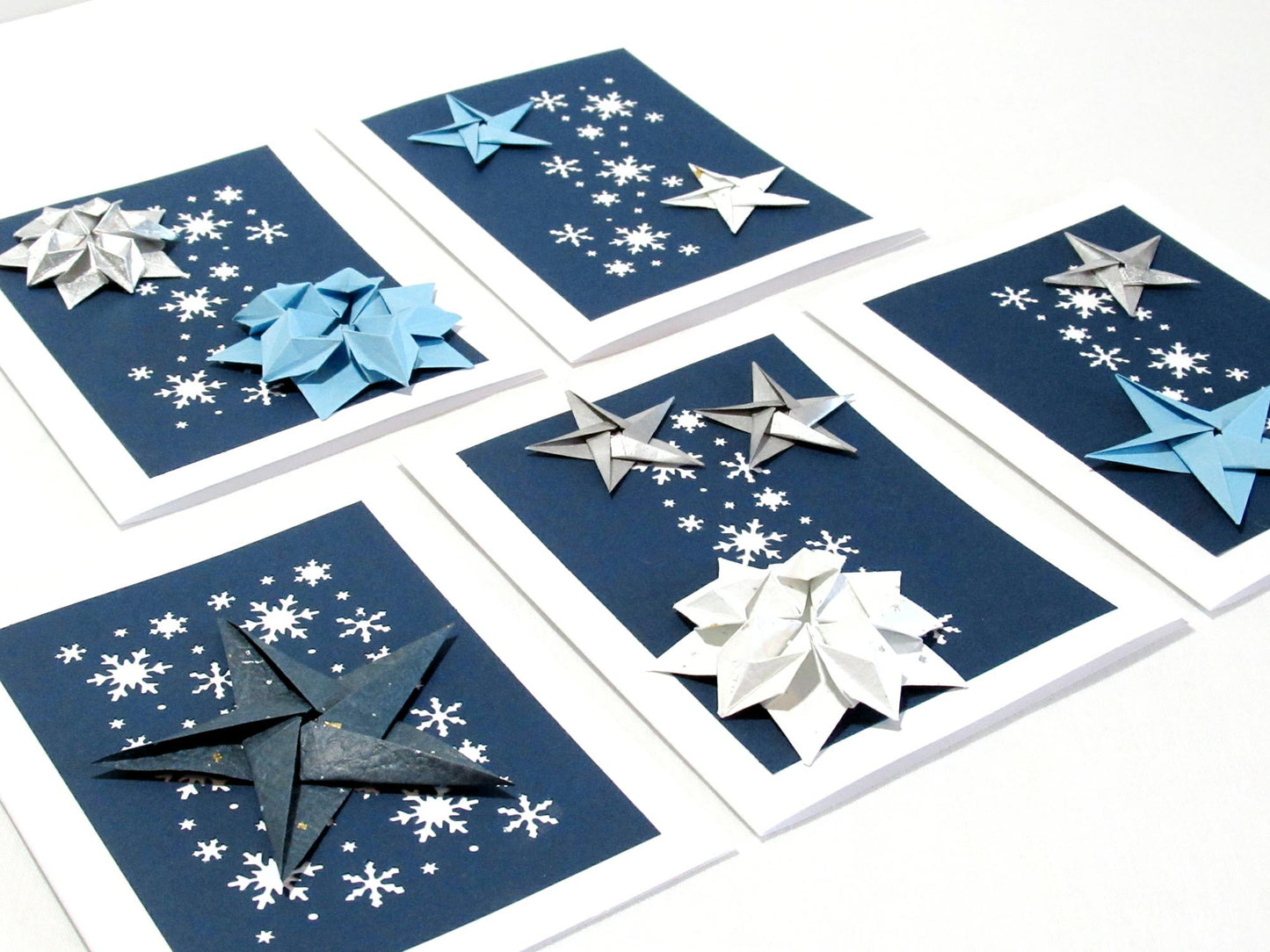 Set of five white cards. Each card has a dark blue background with white snowflakes. On top are a variety of origami stars in white, sliver and blue.