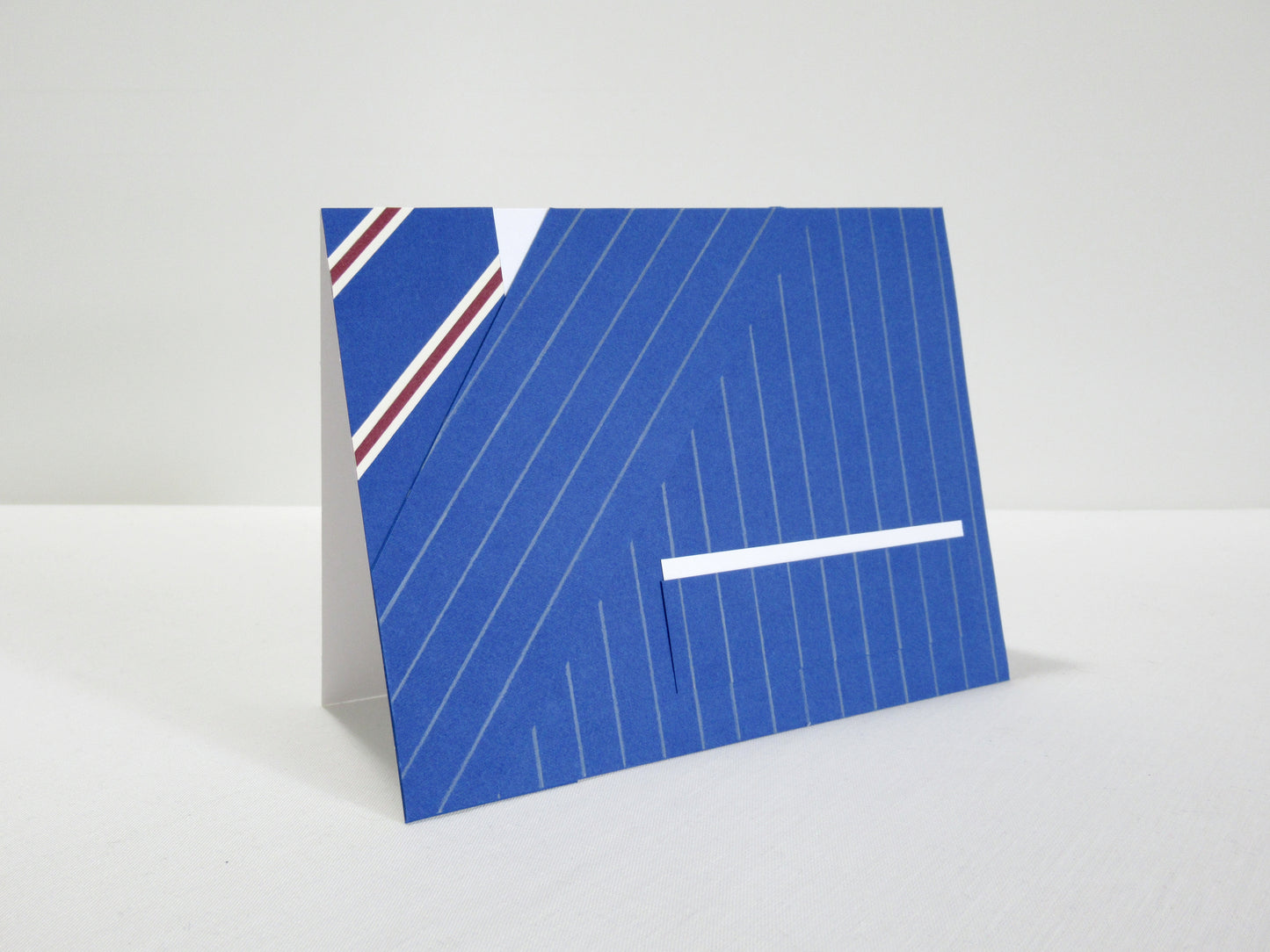 A card stands at an angle on a desk. The front of the card looks like a portion of person's suit, blue pinstripes, white pocket square, and a blue striped tie. It's designed to look like an outfit worn by Harry Hart in the movie Kingsman: The Secret Service.