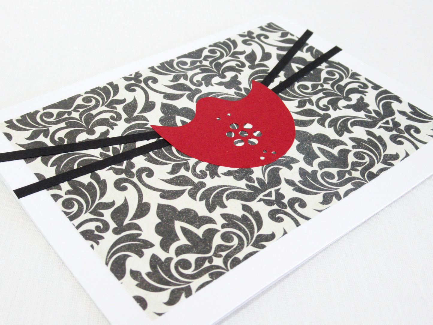 A card lays at an angle on a white desk. The front of the card has an inset with a black and white fillegre background. On top is a red mask that covers just the nose and chin with holes for breathing. Two black straps come out of either side and stretch to the edge of the card. The mask is designed to look like one used in the NBC show Hannibal.