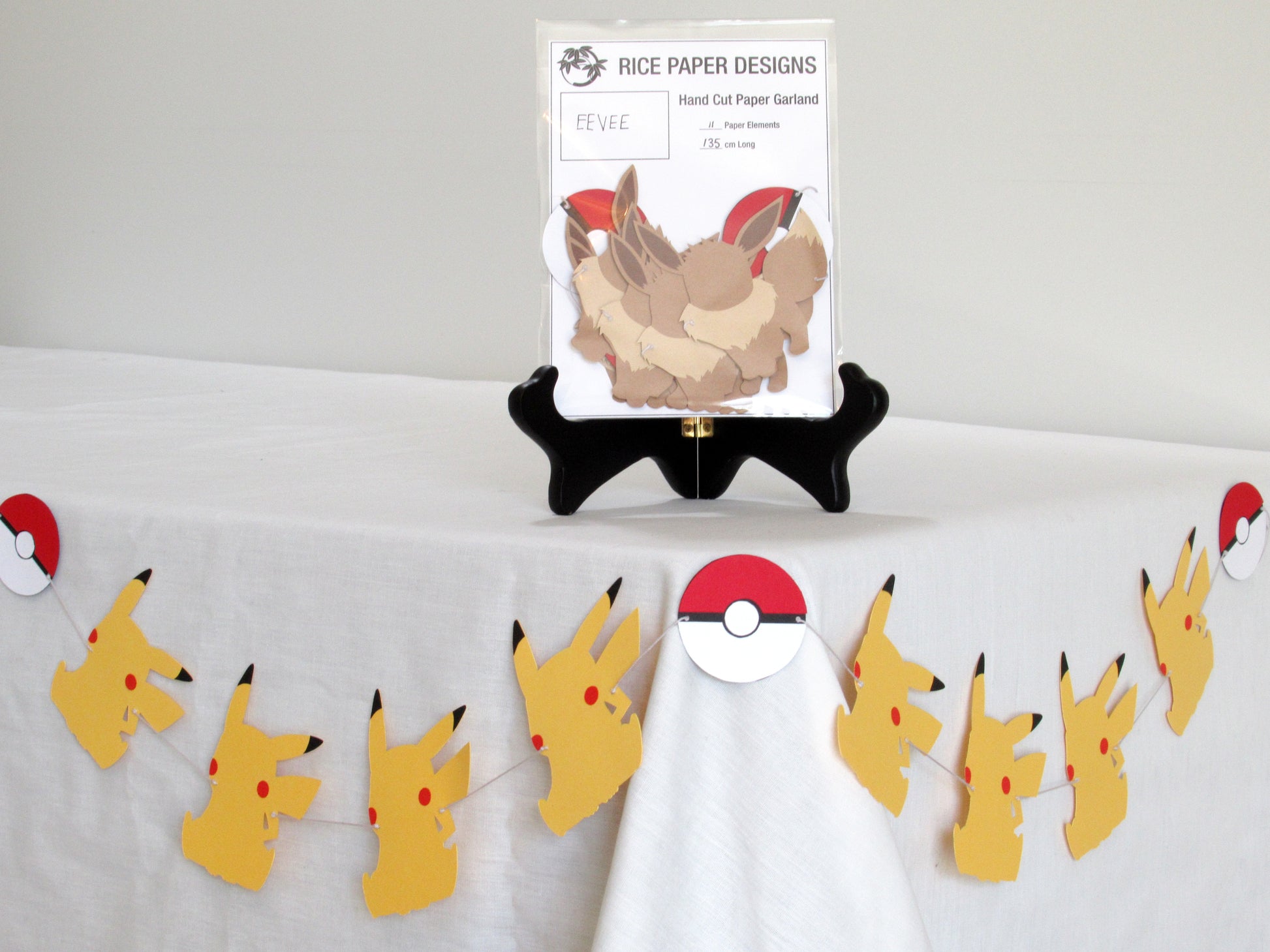 A garland with a series of paper eevee and pokeballs arranged in neat bundle inside a clear bag. There is a paper behind them showing the Rice Paper Designs logo, and information about the garland. The bag is on a table, and below it is a sample garland with pikachu hung up.
