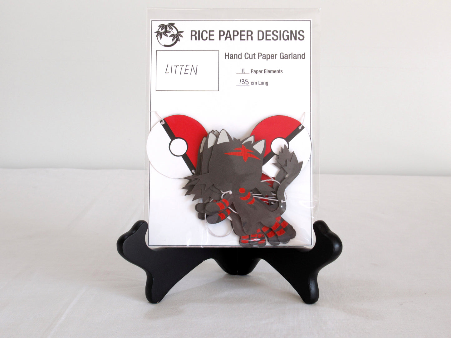 A garland with a series of paper littens and pokeballs arranged in neat bundle inside a clear bag. There is a paper behind them showing the Rice Paper Designs logo, and information about the garland.