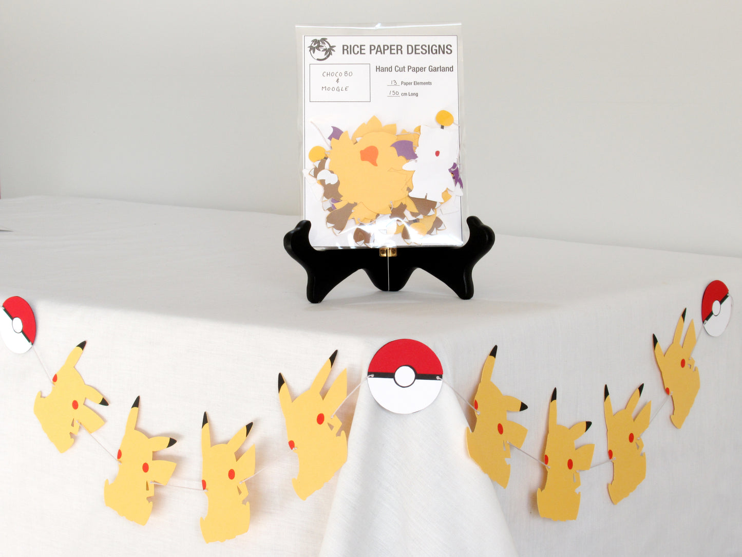 A garland with a series of paper chocobo and moogles arranged in neat bundle inside a clear bag. There is a paper behind them showing the Rice Paper Designs logo, and information about the garland. The bag is on a table, and below it is a sample garland with pikachu hung up.
