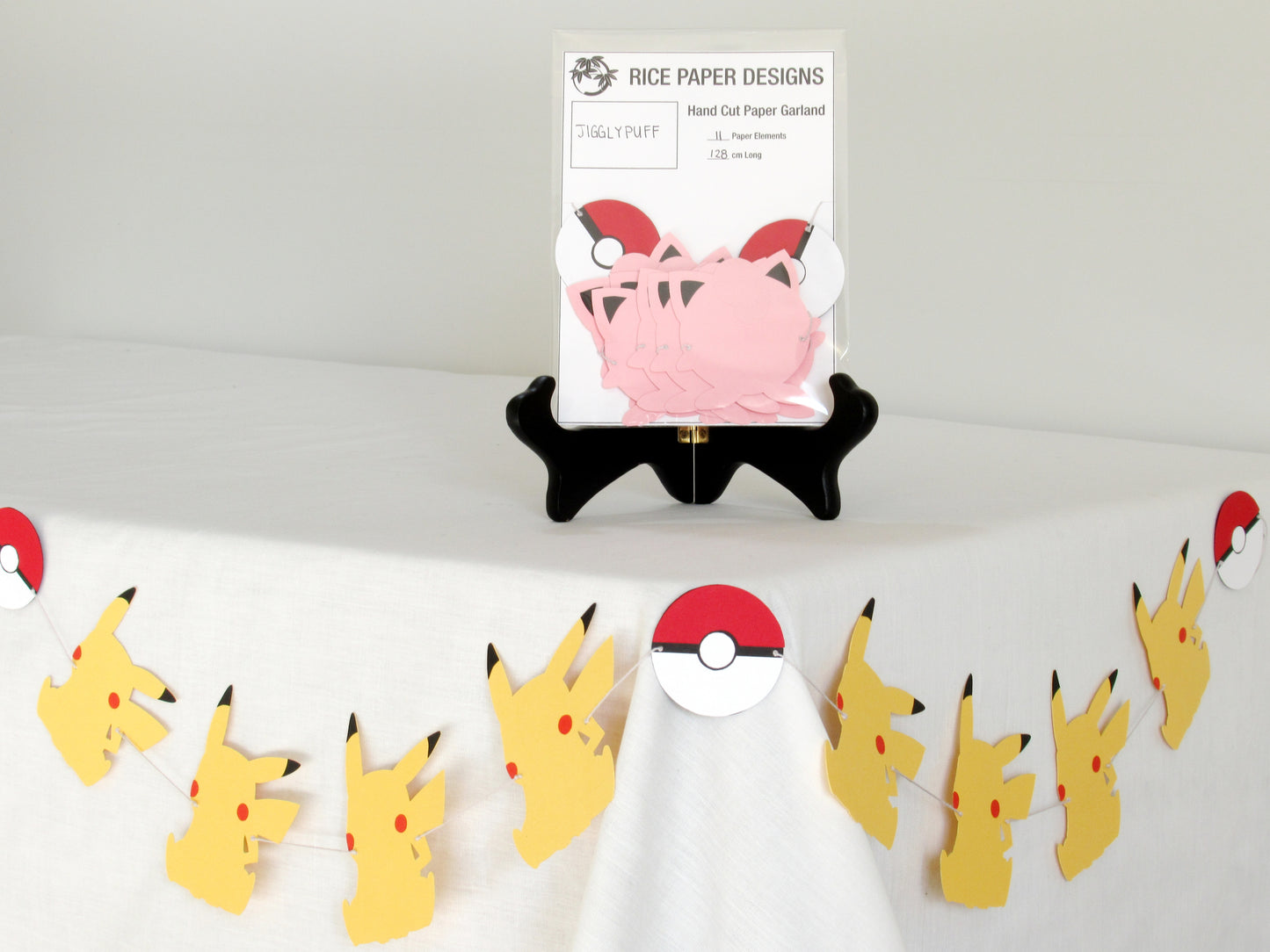 A garland with a series of paper jigglypuffs and pokeballs arranged in neat bundle inside a clear bag. There is a paper behind them showing the Rice Paper Designs logo, and information about the garland. The bag is on a table, and below it is a sample garland with pikachu hung up.