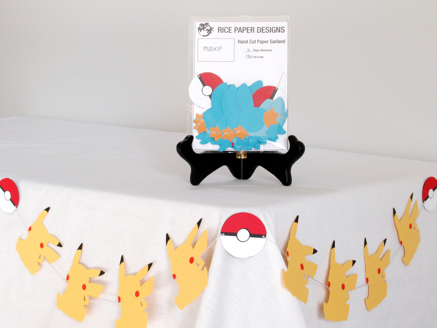 A garland with a series of paper mudkips and pokeballs arranged in neat bundle inside a clear bag. There is a paper behind them showing the Rice Paper Designs logo, and information about the garland. The bag is on a table, and below it is a sample garland with pikachu hung up.