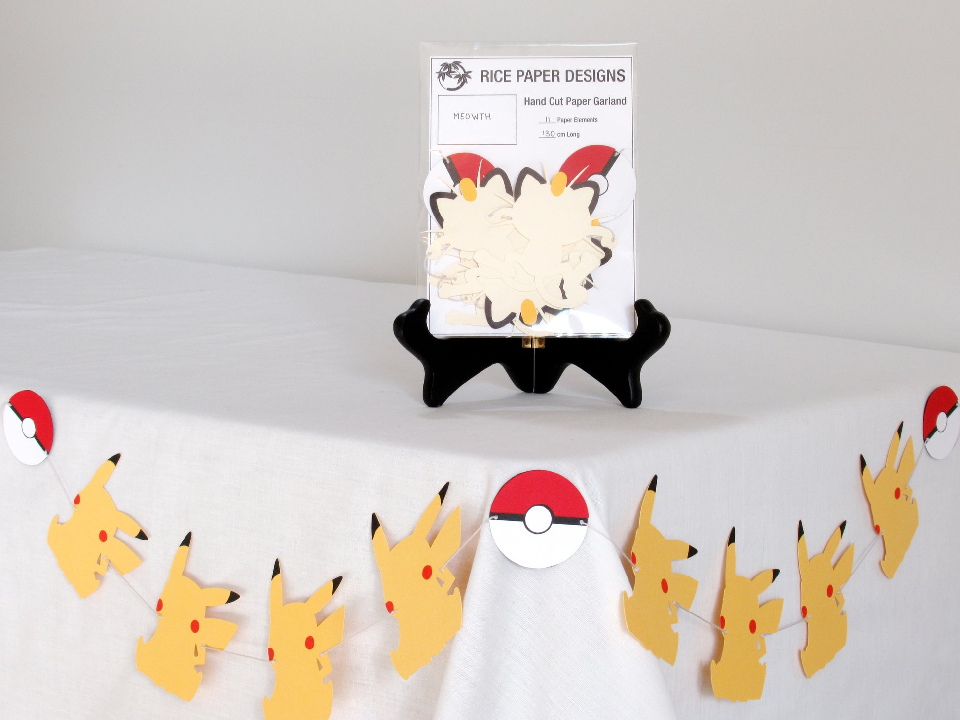 A garland with a series of paper meowths and pokeballs arranged in neat bundle inside a clear bag. There is a paper behind them showing the Rice Paper Designs logo, and information about the garland. The bag is on a table, and below it is a sample garland with pikachu hung up.