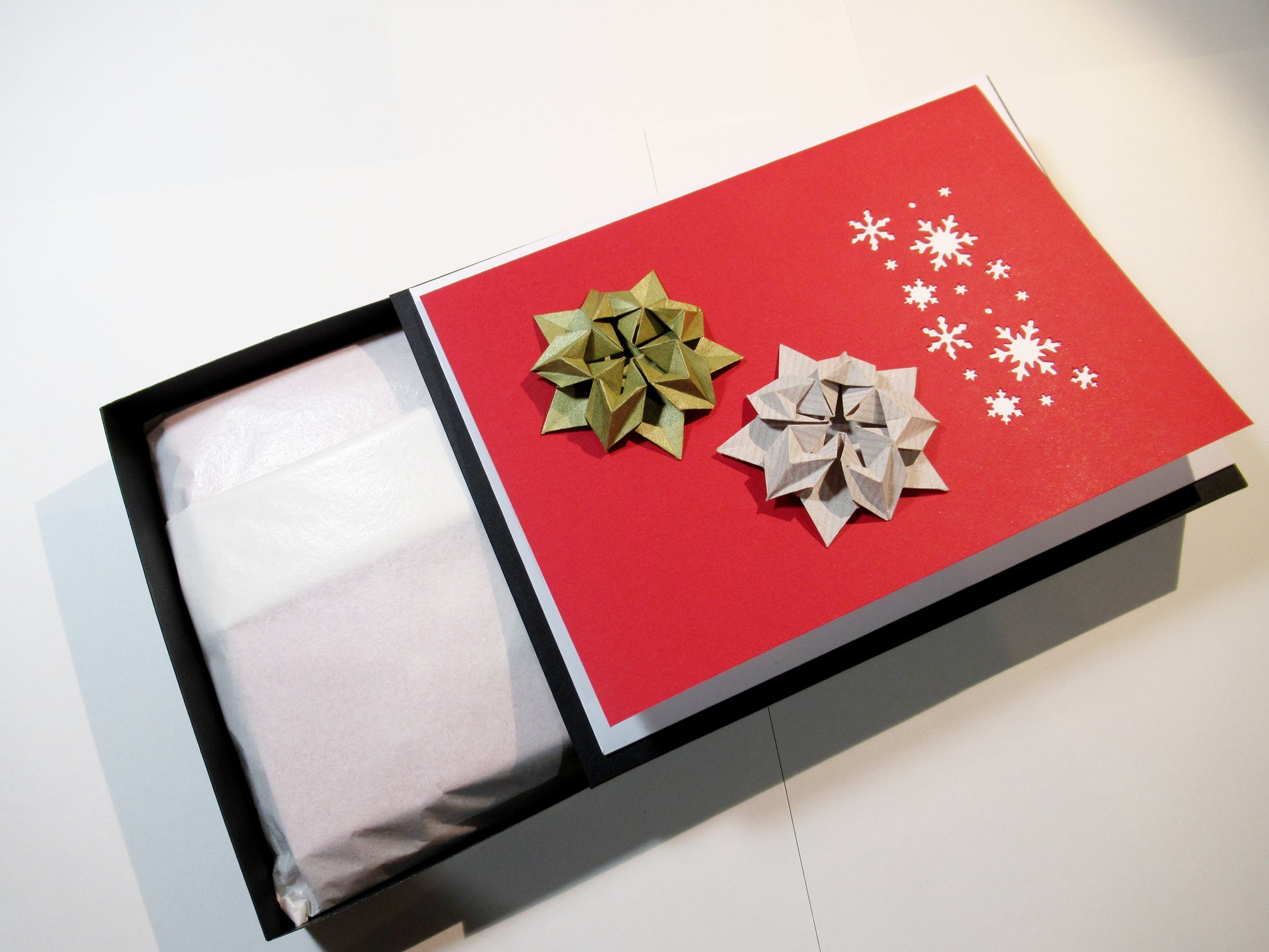 An open black box showing tissue paper wrapped inside. The outside has a red and white card with origami stars and snowflakes resting on top.