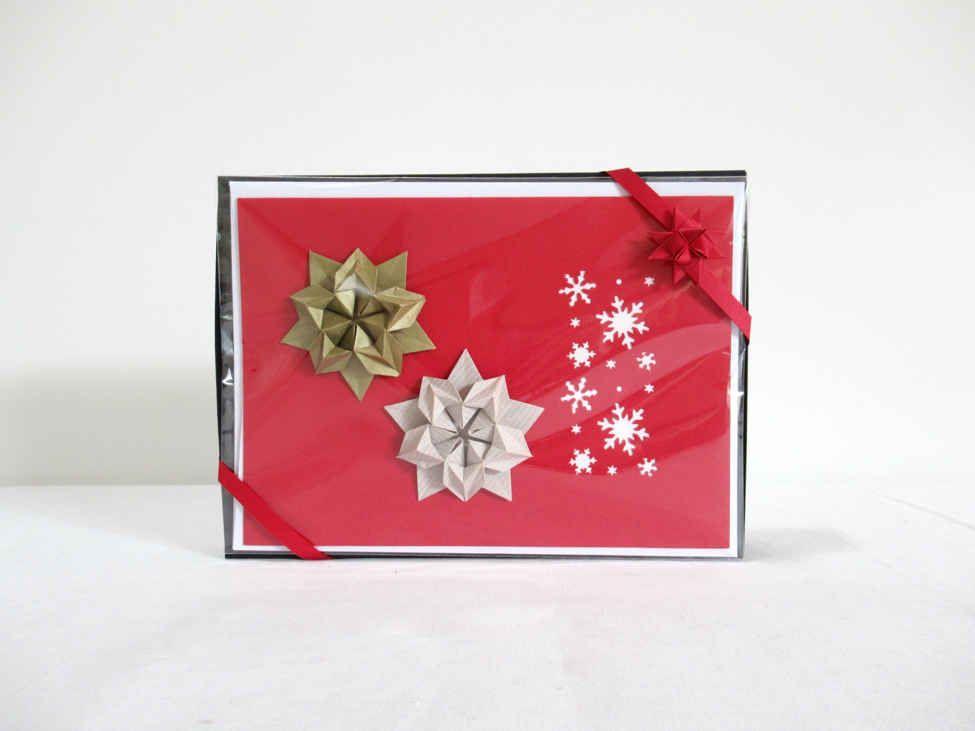 A red and white card, with snowflakes and origami stars, is inside a clear bag. The bag is attached to a black box by two opposite corners, wrapped with a red ribbon with a Froebel star in the corner.