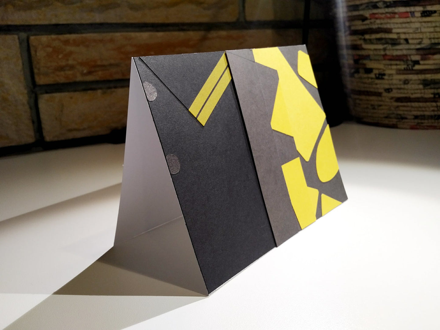A card stands at an angle on a desk. A brown brick wall is in the background. The front of the card looks like a portion of person's shirt, black with yellow details, designed to look like an outfit worn by Eggsy Unwin in the movie Kingsman: The Secret Service.