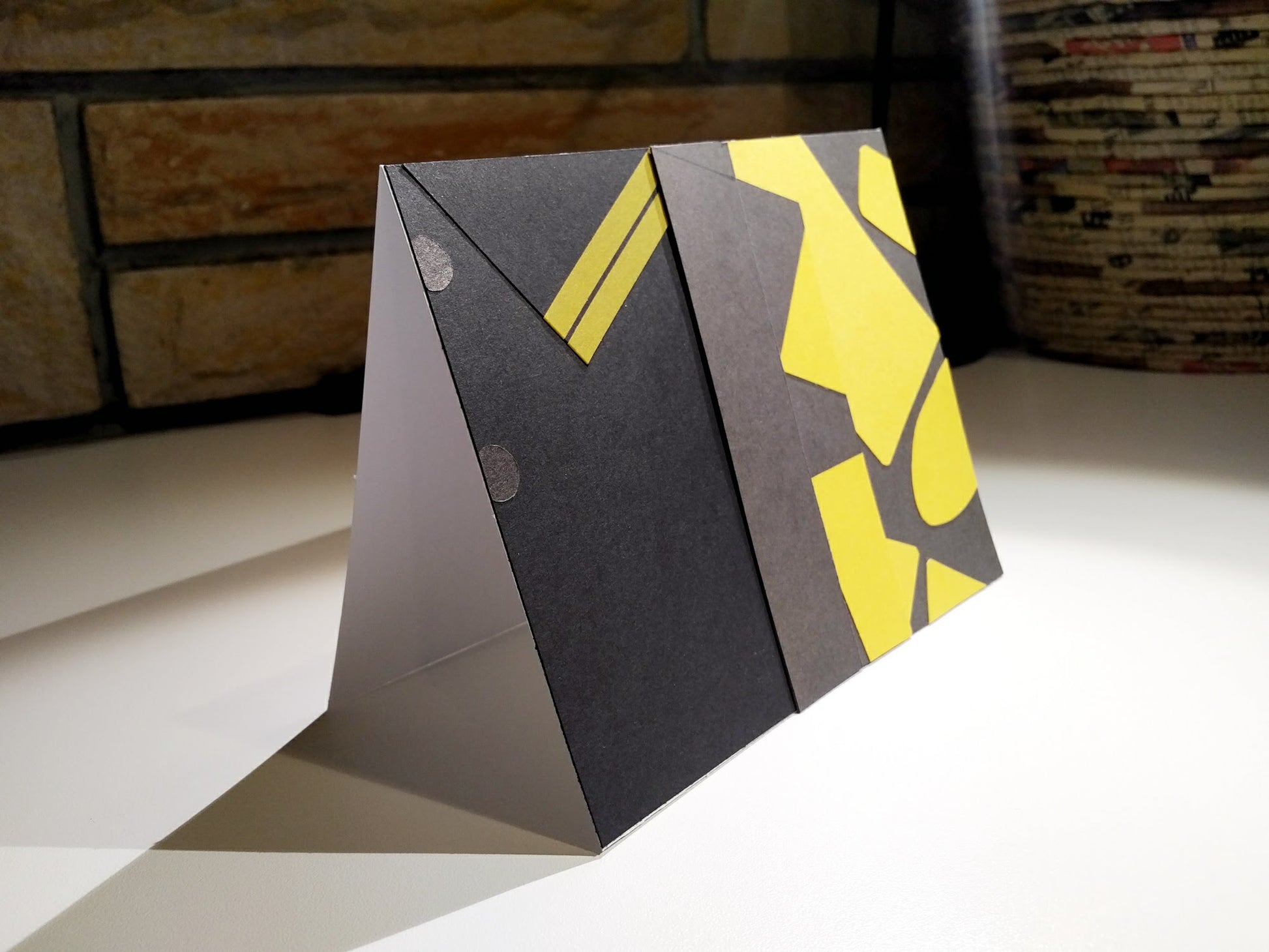 A card stands at an angle on a desk. A brown brick wall is in the background. The front of the card looks like a portion of person's shirt, black with yellow details, designed to look like an outfit worn by Eggsy Unwin in the movie Kingsman: The Secret Service.