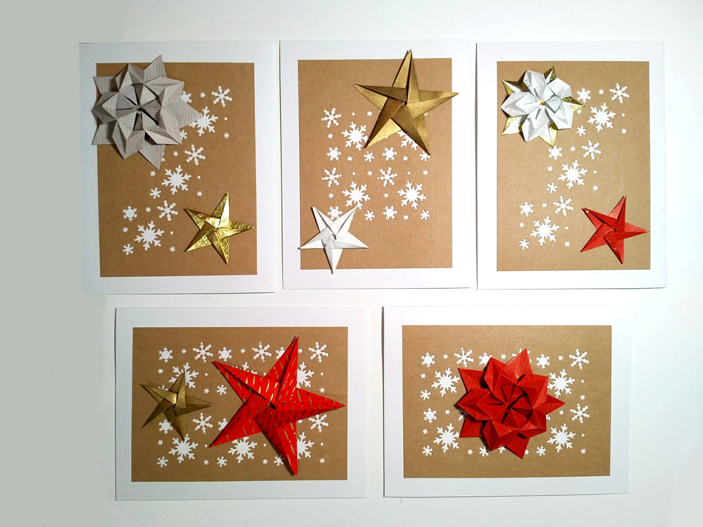 Set of five white cards. Each card has a kraft background with white snowflakes. On top are a variety of origami stars in white, gold and red.