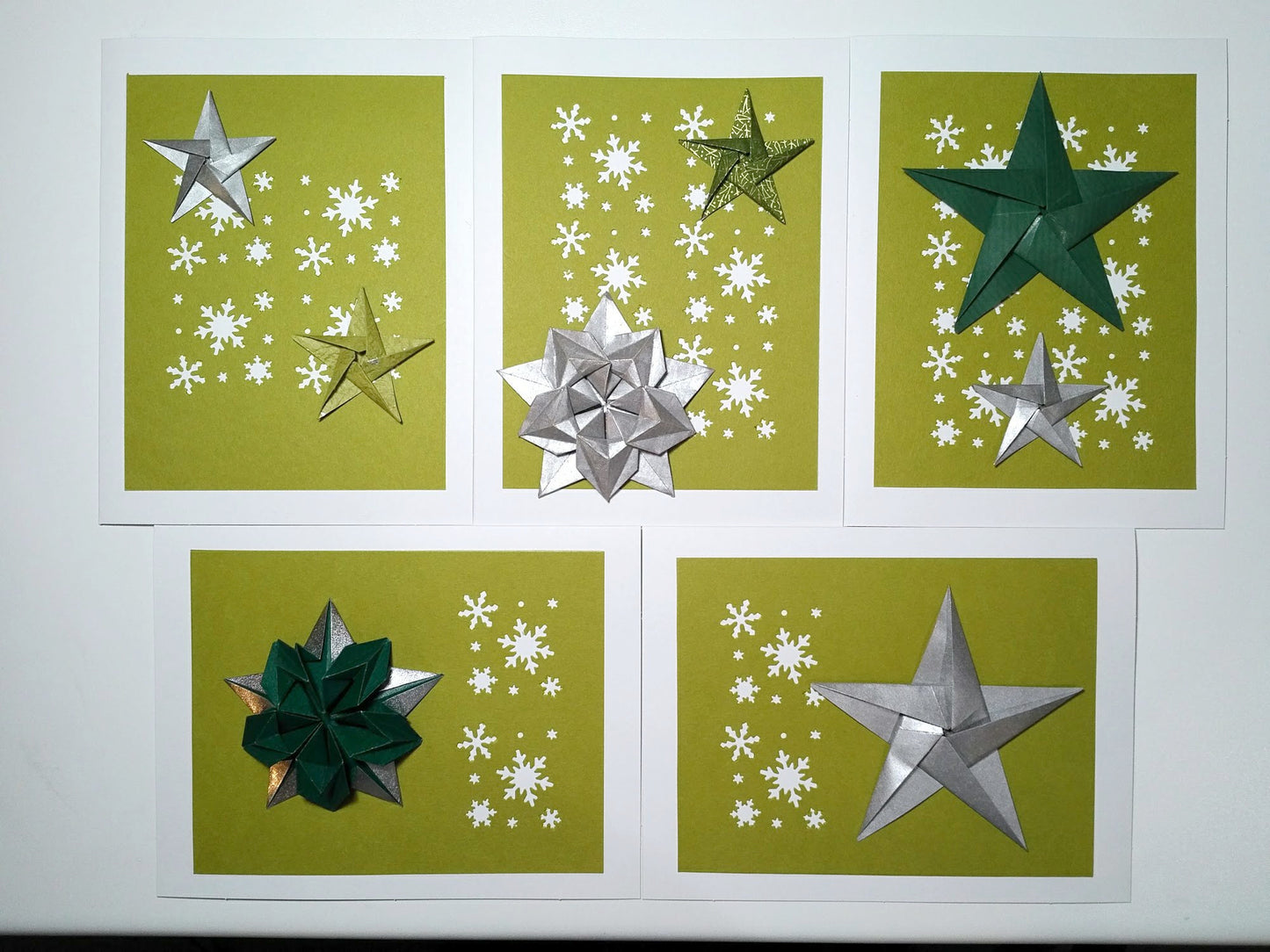 Set of five white cards. Each card has a green background with white snowflakes. On top are a variety of origami stars in both silver and green.