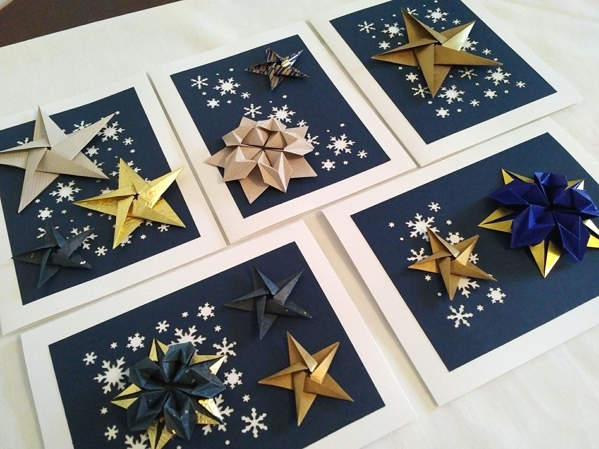 Set of five white cards. Each card has a dark blue background with white snowflakes. On top are a variety of origami stars in white, gold and blue.
