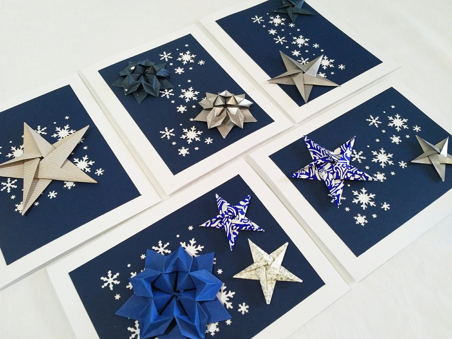 Set of five white cards. Each card has a dark blue background with white snowflakes. On top are a variety of origami stars in silver, white, and blue.