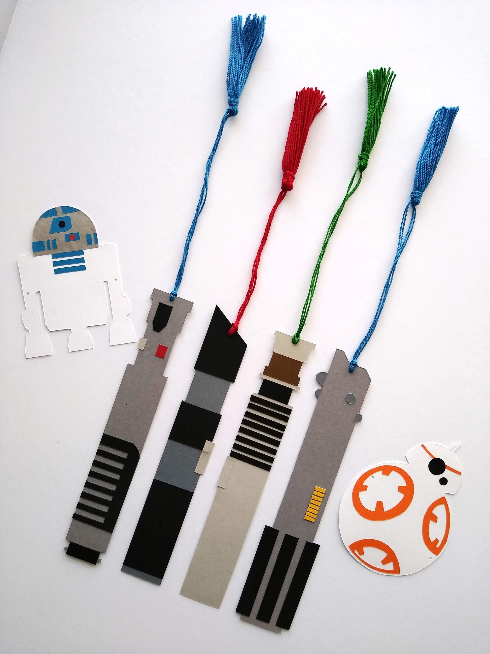 Four bookmarks with tassels stretched out lay on a white background. They rest between a paper R2D2 and BB8. Each bookmark is designed to look like a light saber from Star Wars, with the light portion represented by a colored tassel.