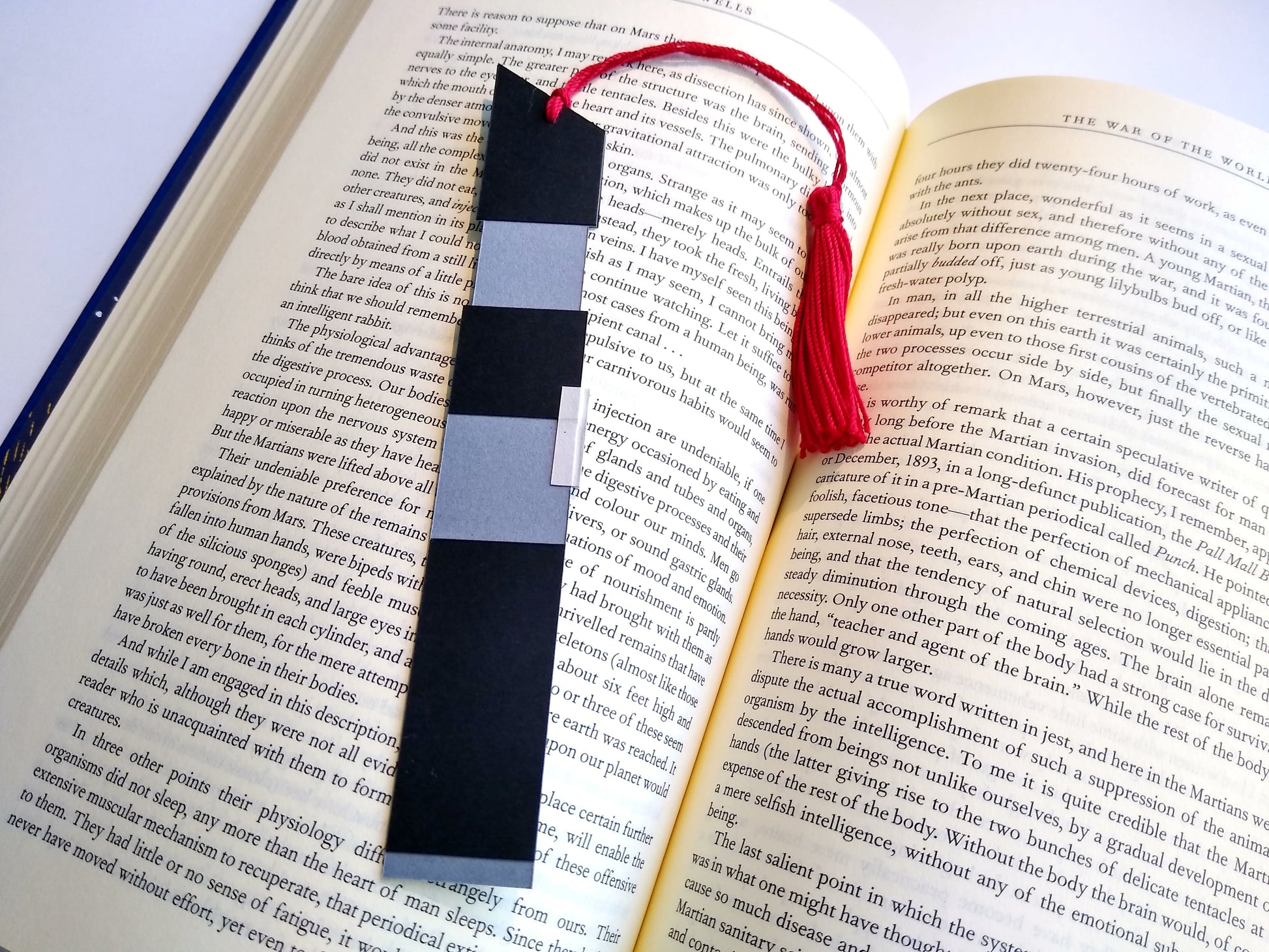 A bookmark designed like the handle of Darth Vader's lightsaber rests on top of an open book. The red tassel curls around and rests in dip between the pages.