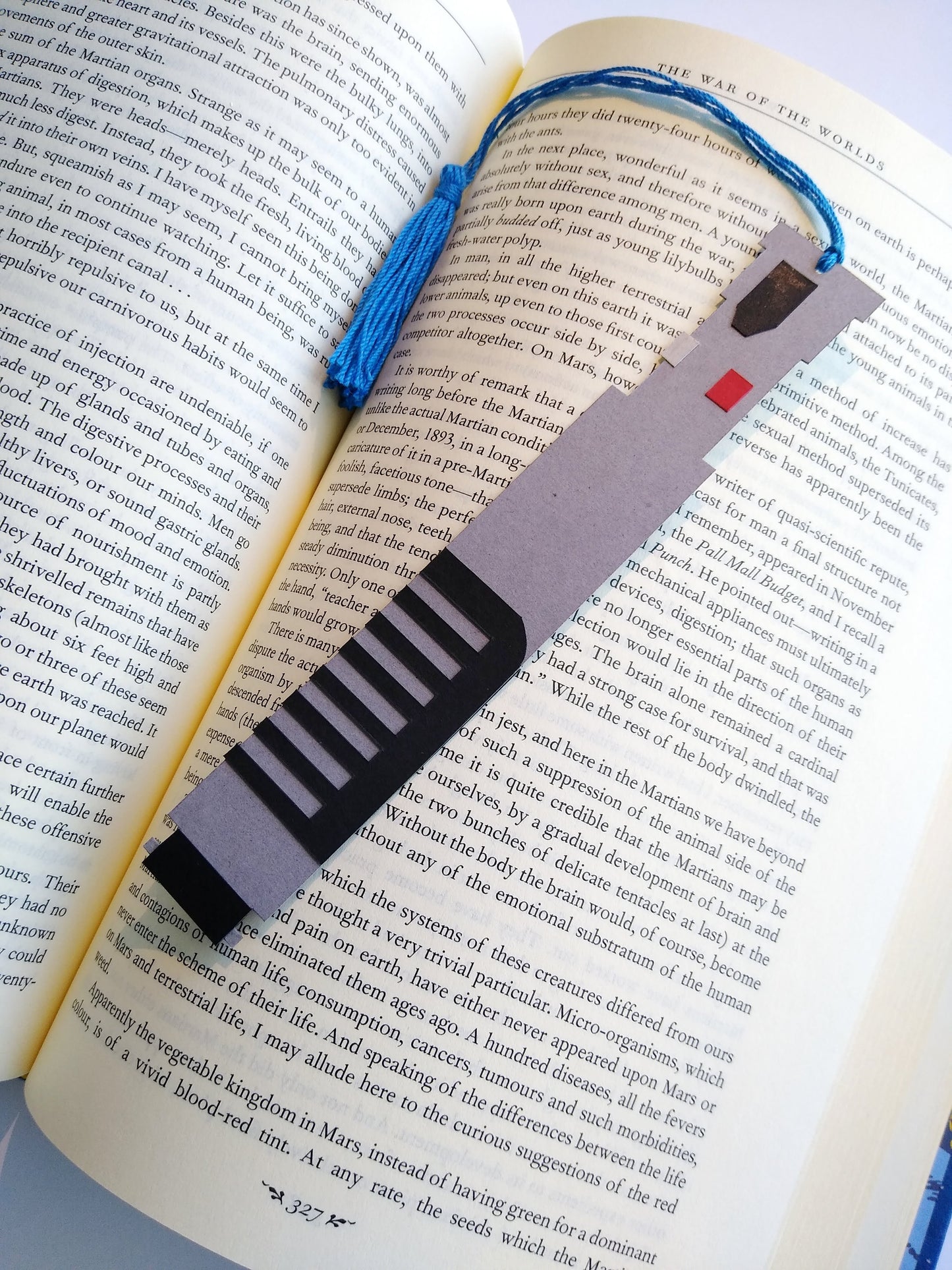 A bookmark designed like the handle of Obi-Wan Kenobi's lightsaber rests on top of an open book. The blue tassel curls around and rests in dip between the pages.