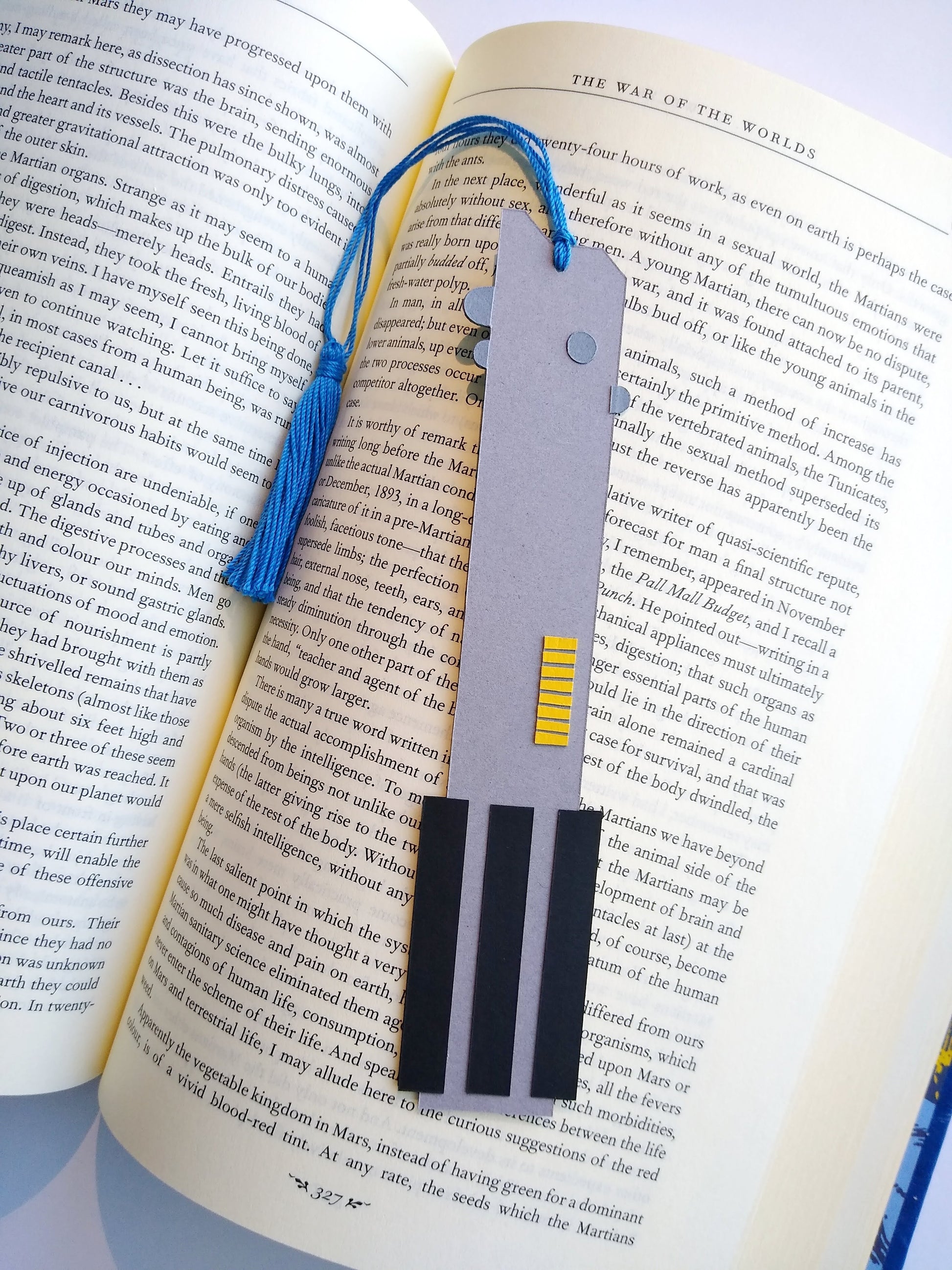 A bookmark designed like the handle of Anakin Skywalker's lightsaber rests on top of an open book. The blue tassel curls around and rests in dip between the pages.