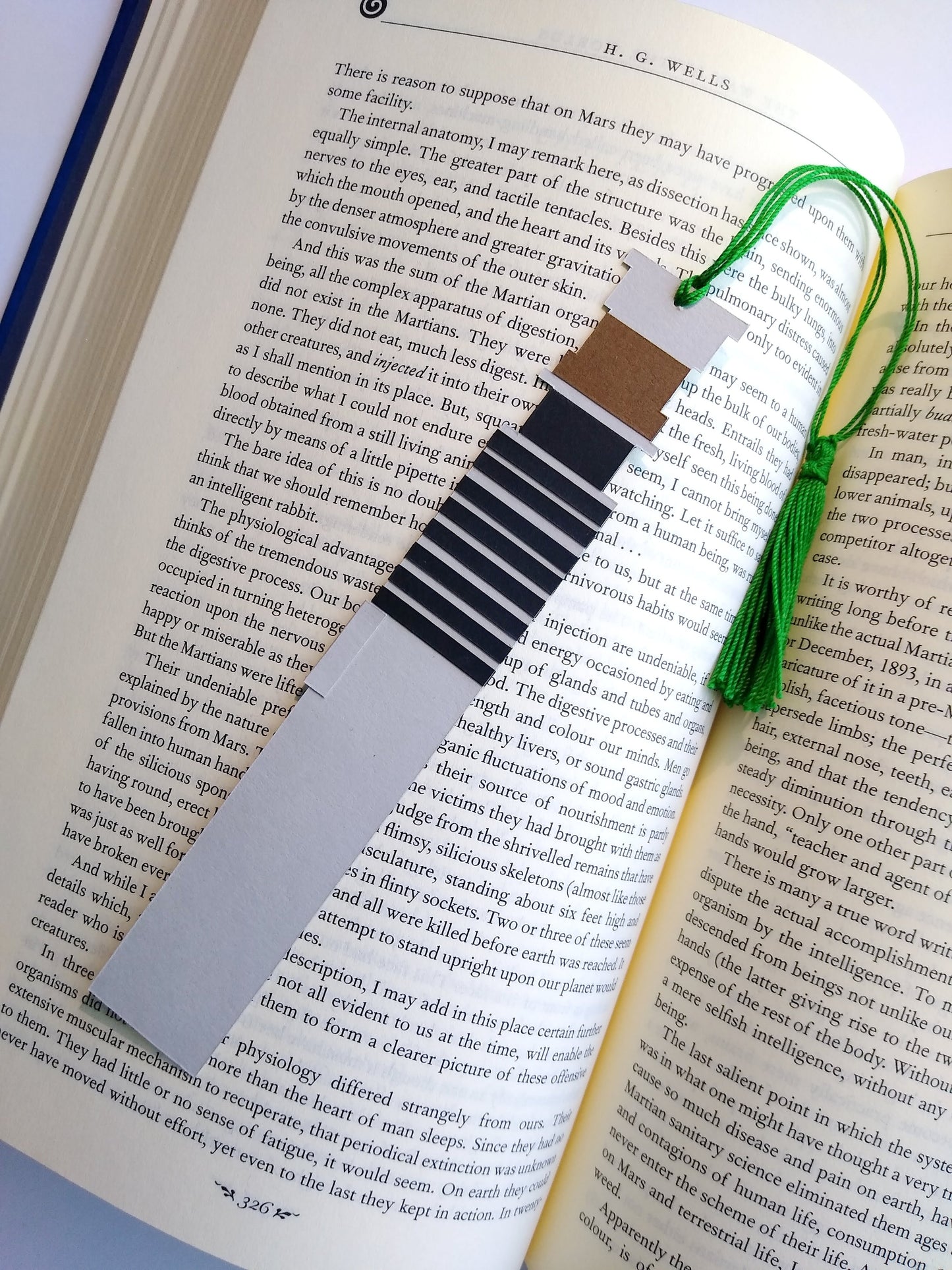 A bookmark designed like the handle of Luke Skywalker's lightsaber rests on top of an open book. The green tassel curls around and rests in dip between the pages.