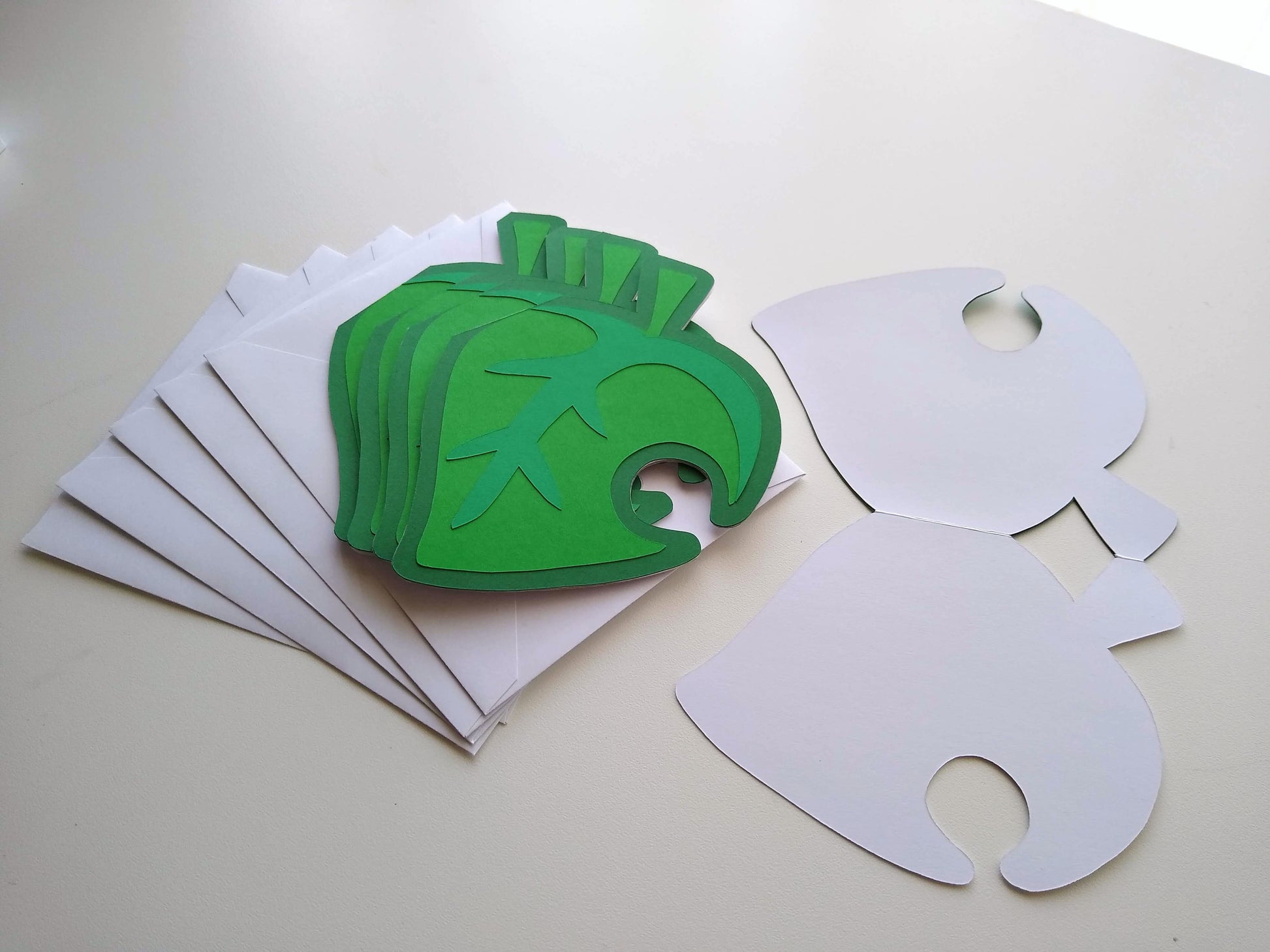 Four cards designed to look like green leaves with a hole in the side are stacked, splayed, on top of five white envelopes. One card is opened up flat on the table beside them.