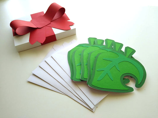 Five cards  designed to look like green leaves with a hole in the side are stacked, splayed, on top of five white envelopes. The cards and envelopes are next to a white box with a big red, paper ribbon on it made to look like a wrapped package.