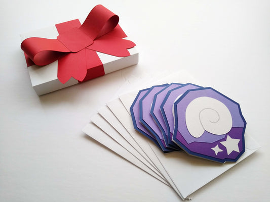 Five purple and white cards are stacked, splayed, on top of five white envelopes. The cards are designed to look like a shell and stars stuck inside a purple rock, like fossils. The cards and envelopes are next to a white box with a big red, paper ribbon on it made to look like a wrapped package.